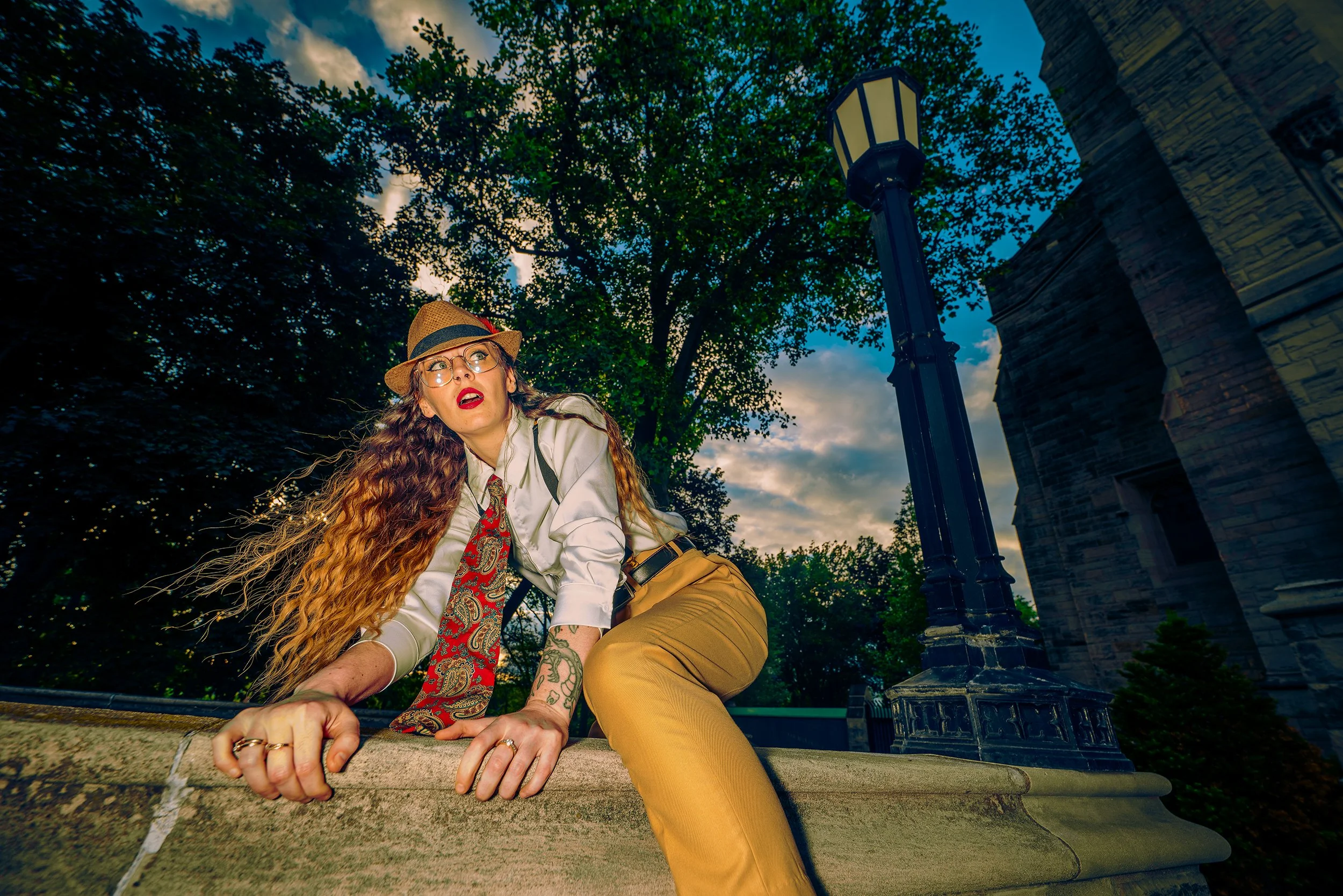 Woman with long curly hair, glasses, red lipstick, wearing a hat, white shirt, paisley tie, and yellow pants crawling on a ledge outdoors near a brick building and lamp post during sunset.