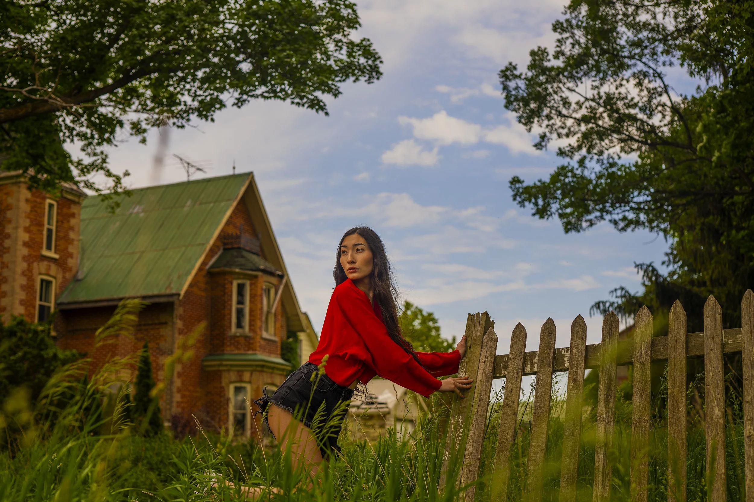A young woman in a red jacket and black shorts leaning on a wooden fence in a yard with tall grass, with a large brick house with a green roof and trees in the background, under a partly cloudy sky.