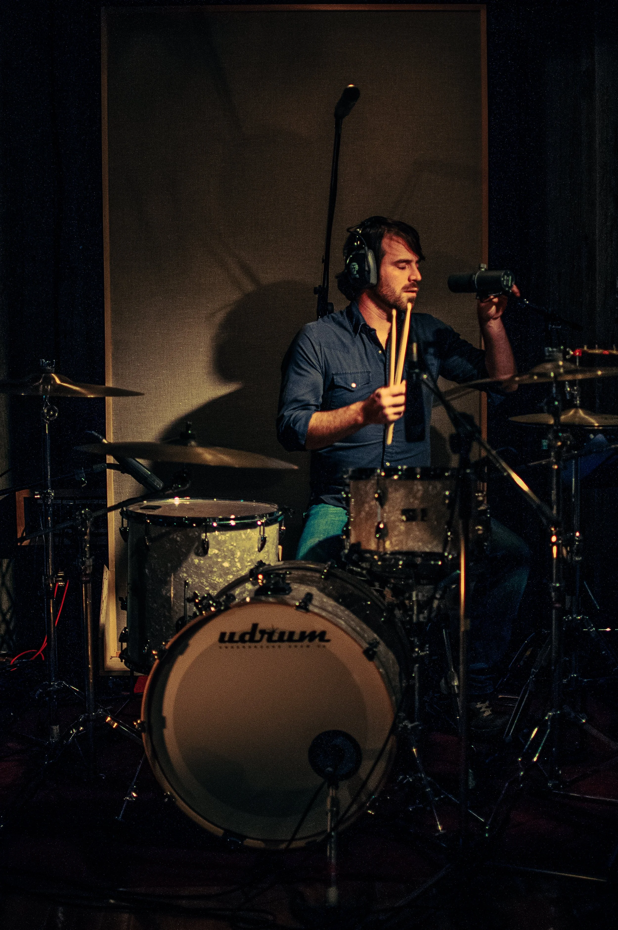 A man wearing headphones and a dark shirt is playing a drum set in a recording studio, with a microphone set up in front of him. The background is dark, and the drum set includes a bass drum and cymbals.