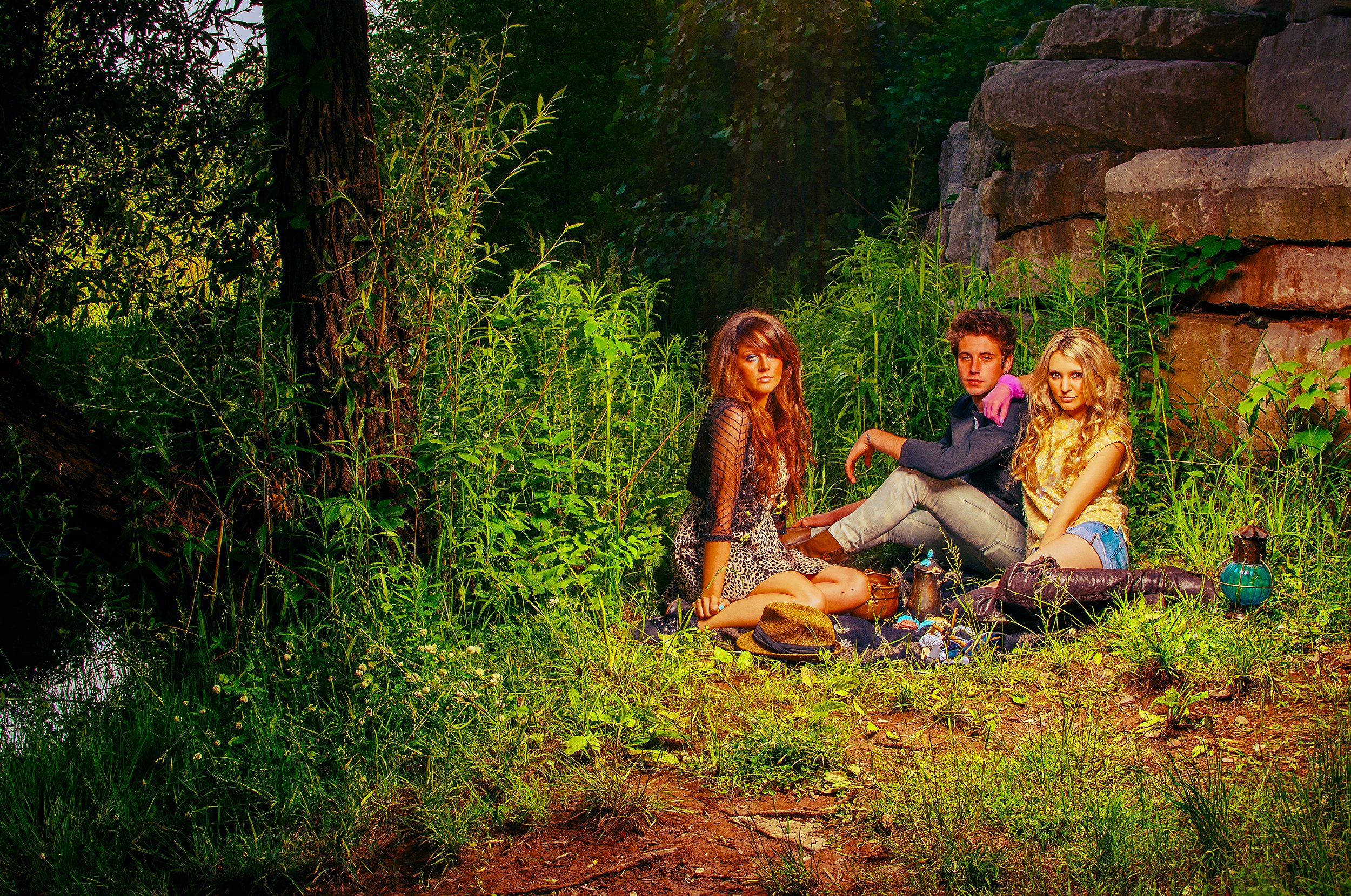 Three young adults, two women and one man, sitting and lying on the grass in a lush forest with tall trees and rocks. They are surrounded by greenery and outdoor camping gear.