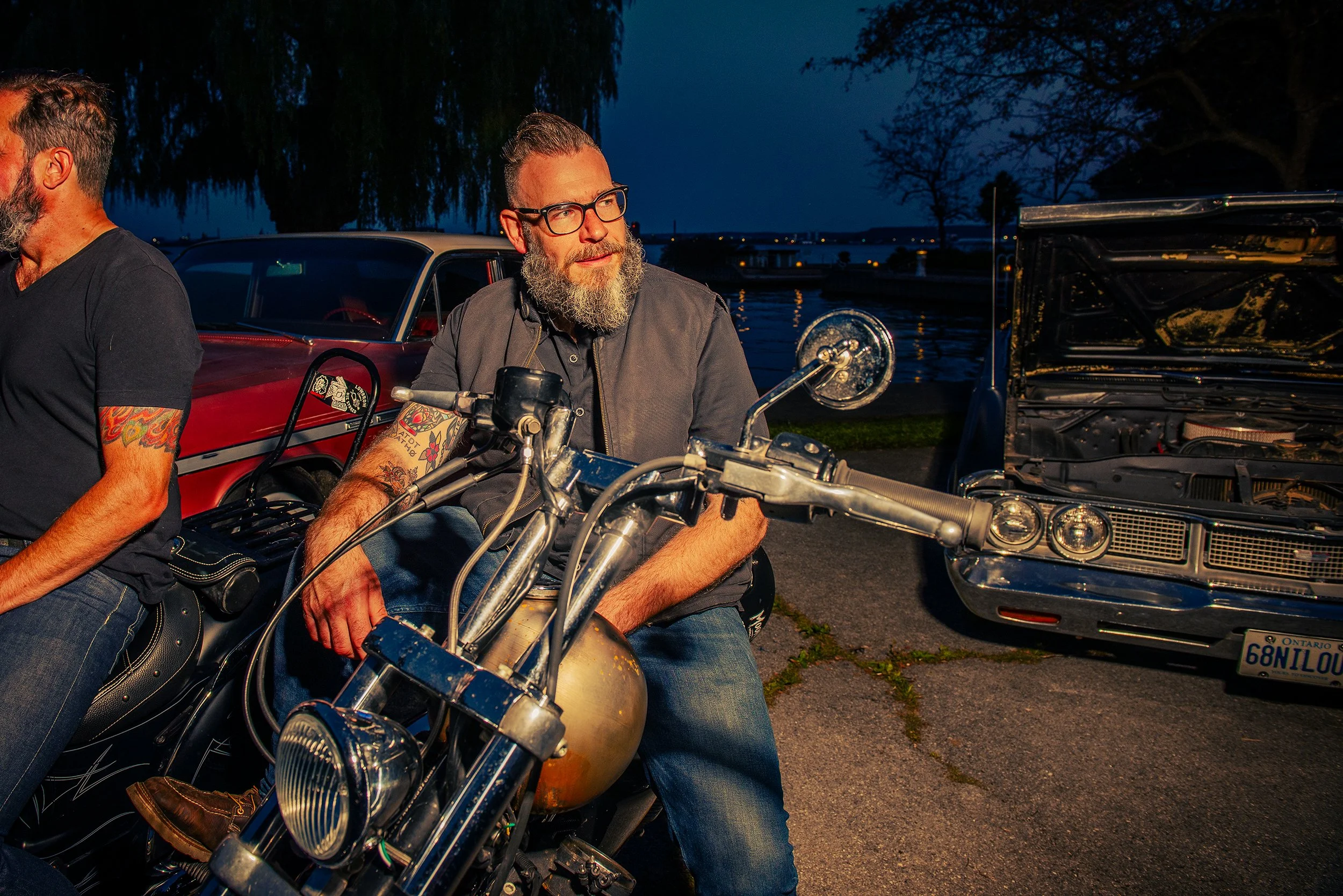 Two men sitting on motorcycles near parked cars along a waterfront at dusk, with trees and water in the background.