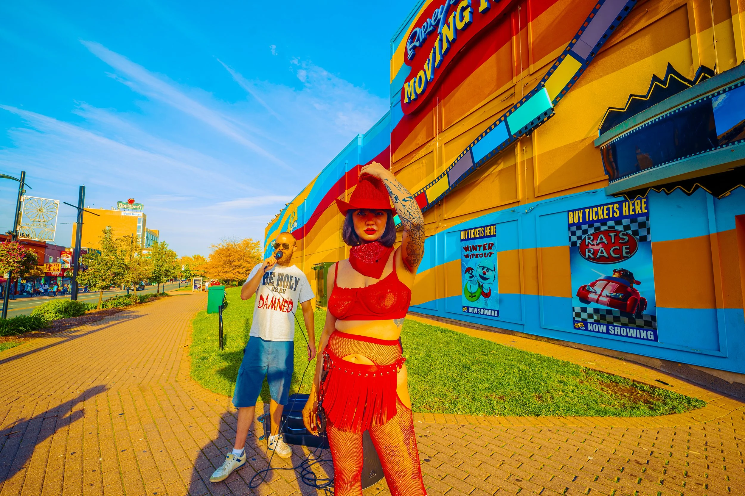 A woman in a red outfit, hat, and fishnet stockings posing in front of a colorful amusement park or carnival building with a sign for a rap race. A man next to her is singing or speaking into a microphone. The sky is bright blue with a few clouds, an