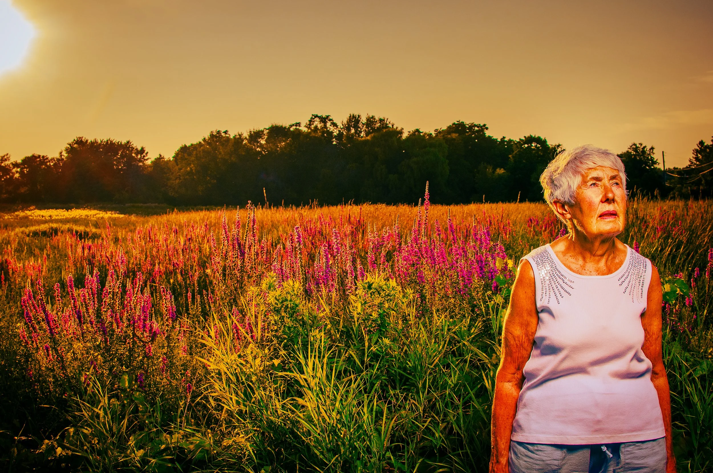 An elderly woman with gray hair wearing a white sleeveless top and gray shorts stands in a field of pink and purple flowers during sunset, with trees in the background.