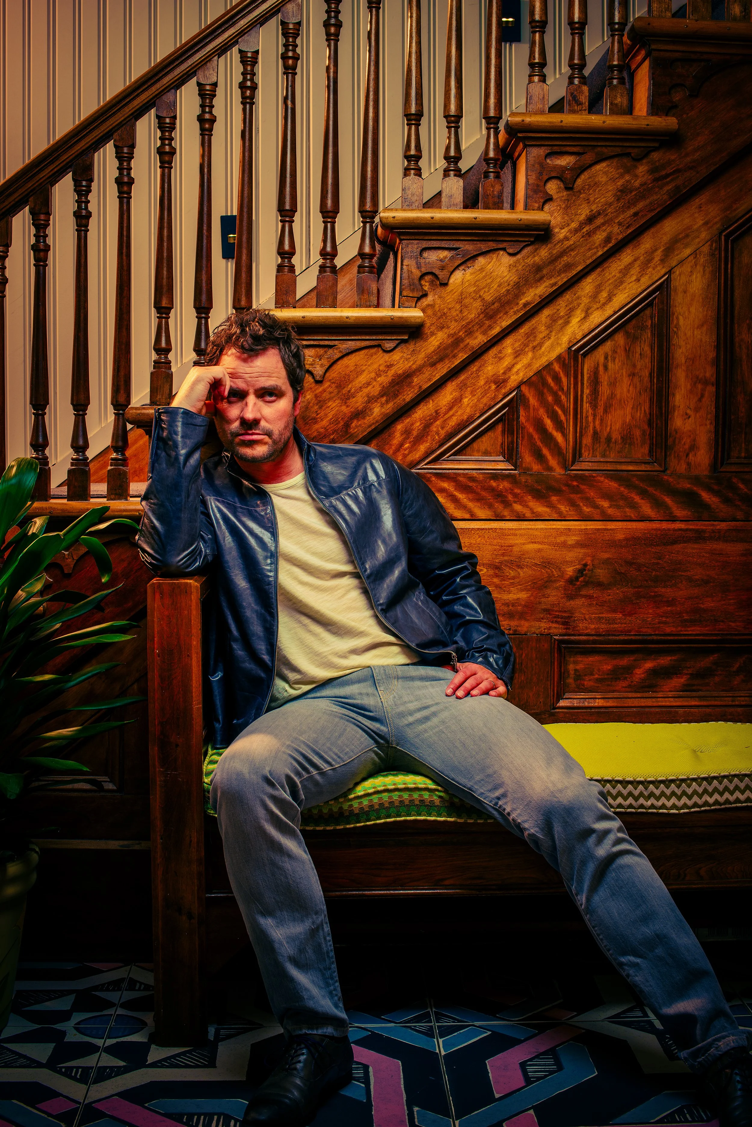 A man with curly dark hair and a beard, wearing a black leather jacket, sitting on a colorful bench in front of a wooden staircase with spindled balusters. The man is resting his head on his left hand, looking directly at the camera, and appears to b