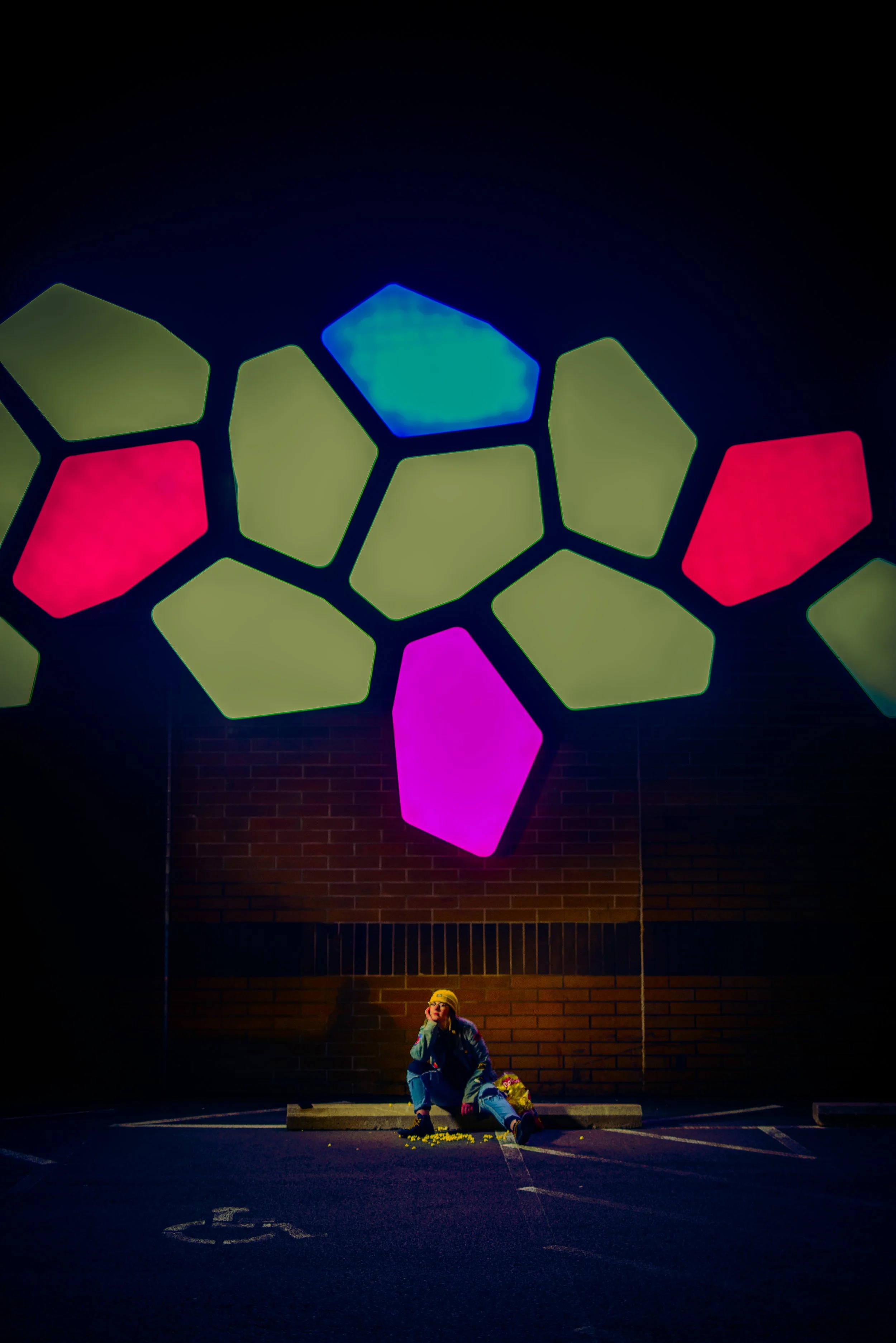 A person sitting on the ground beneath a colorful illuminated tree installation in a dark outdoor setting.