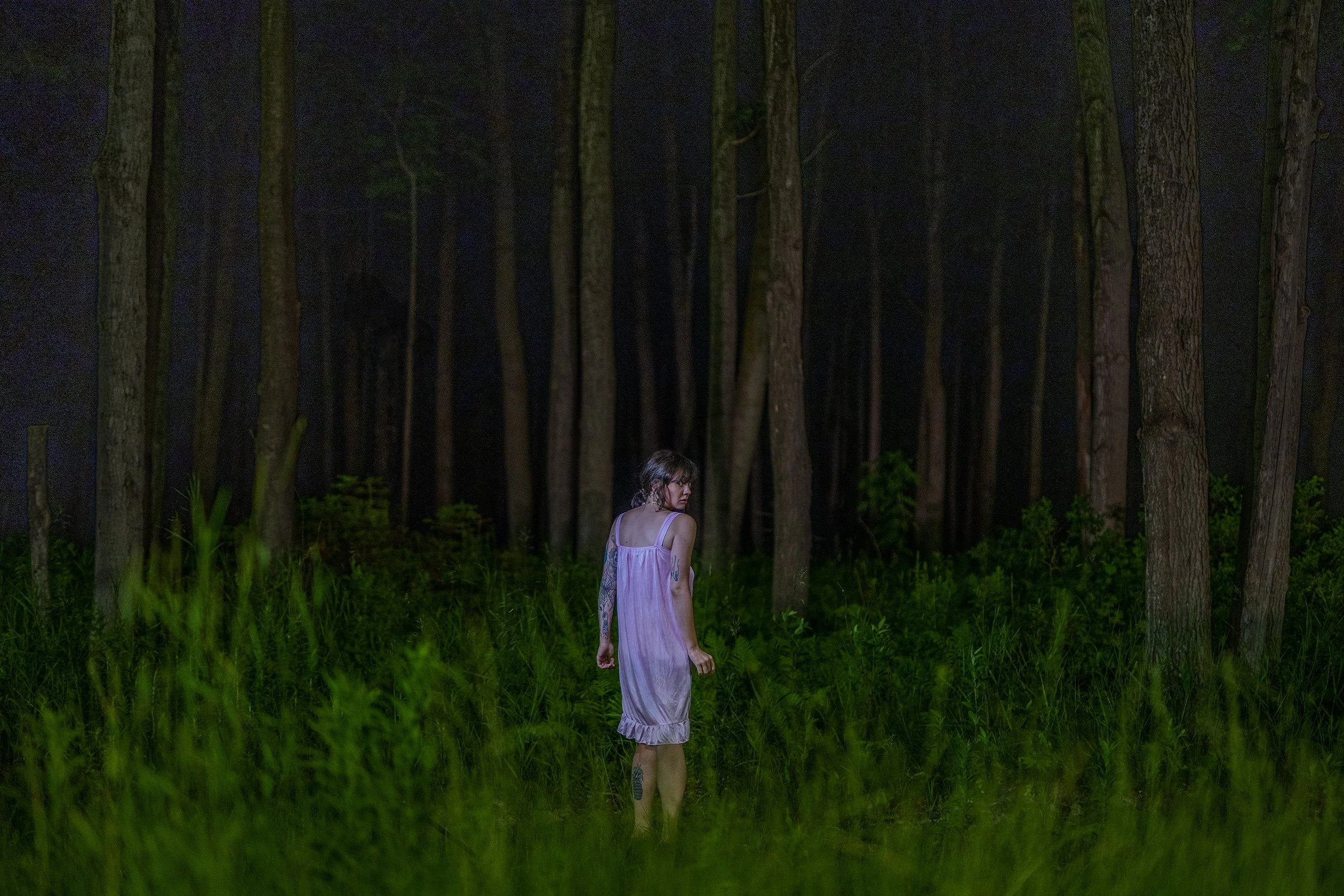 A woman with dark hair and tattoos wearing a pink dress standing in a dark forest at night.