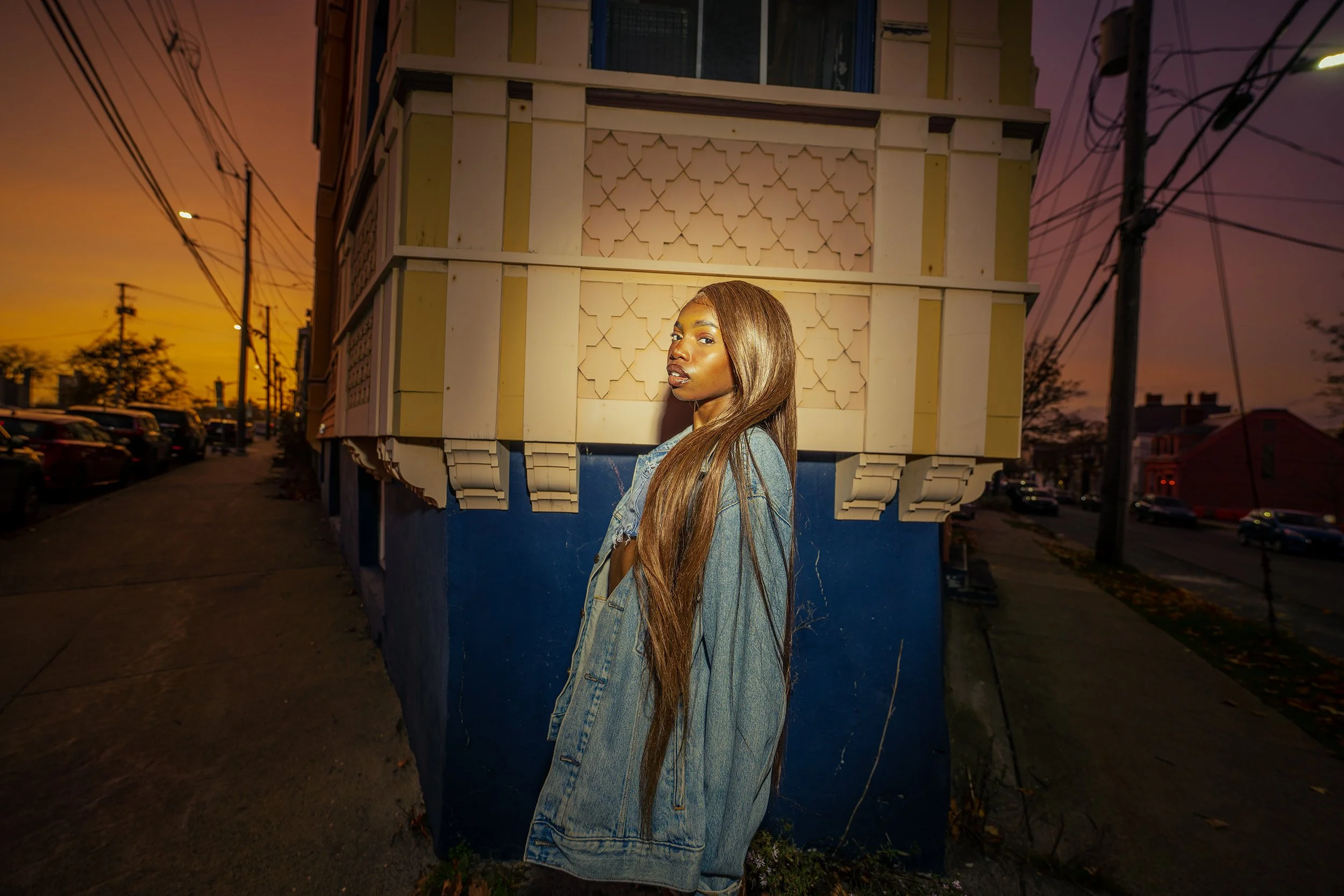 A young woman with long brown hair wearing a denim jacket standing against a building wall at sunset.