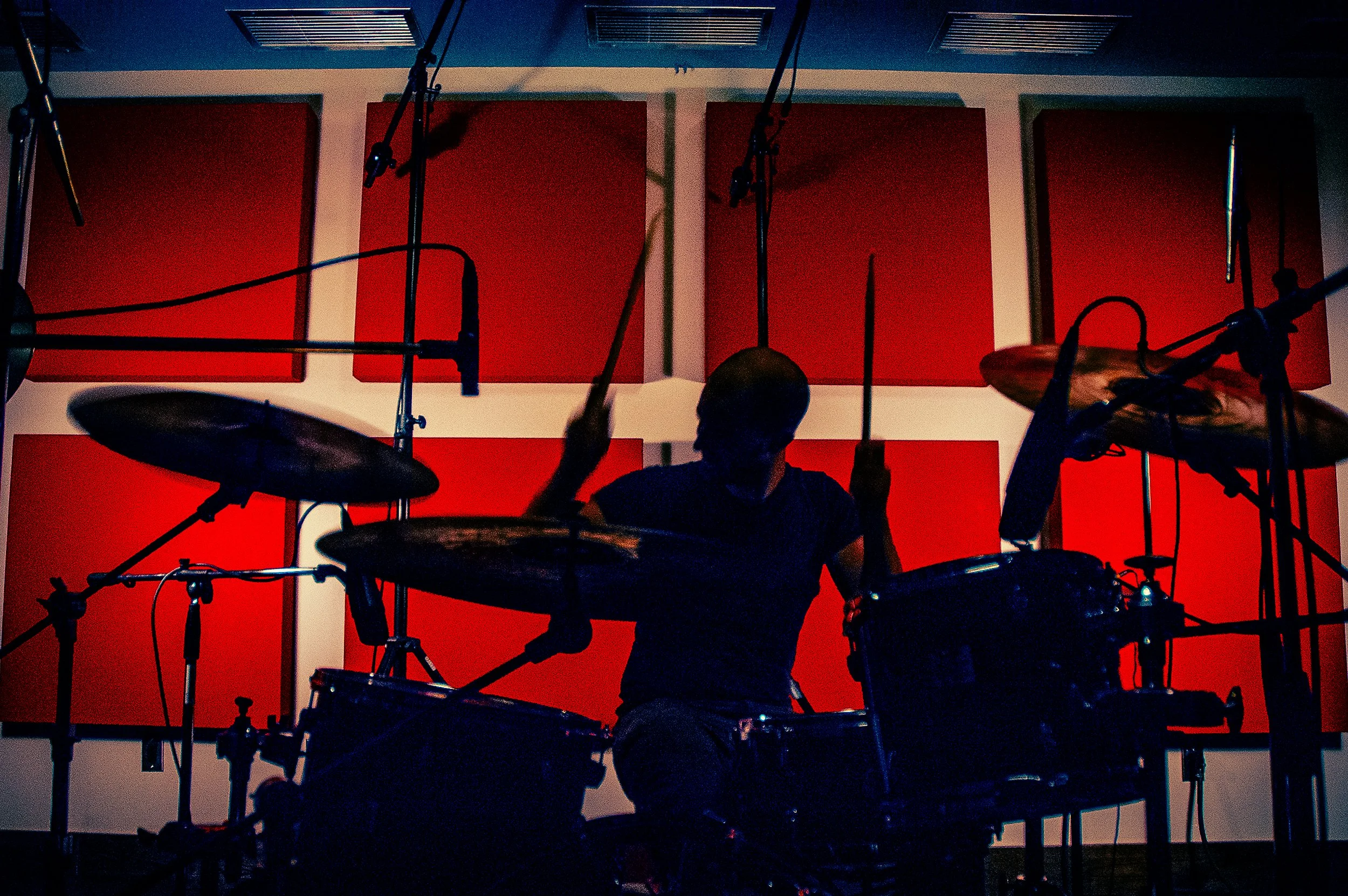 A silhouette of a drummer playing a drum set in a recording studio with red soundproof panels on the wall behind.
