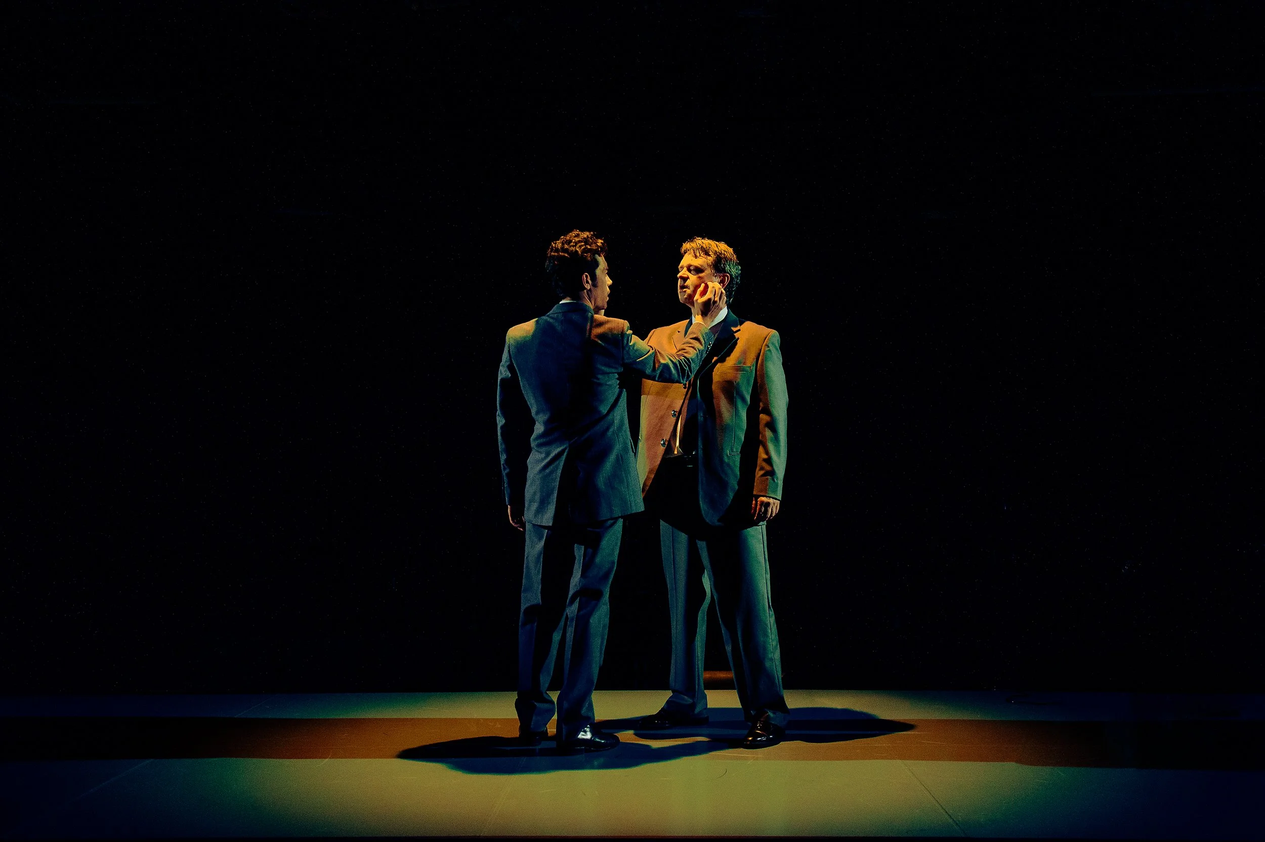 Two men dressed in suits are standing on a dark stage with a black background, engaged in an intense emotional confrontation, with one man touching the other's face.