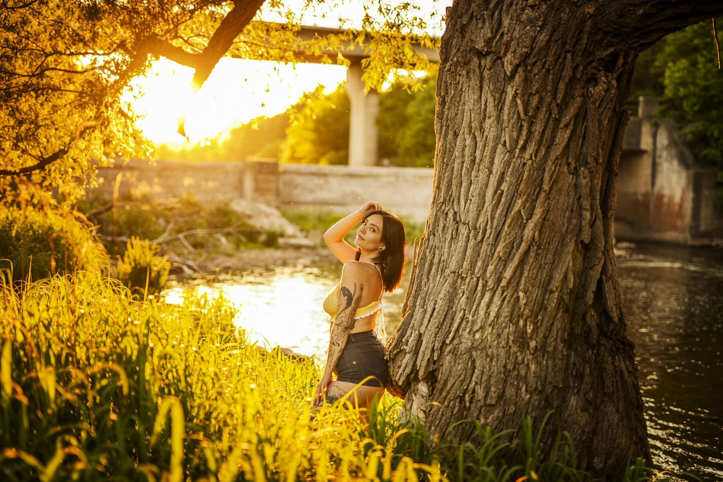 A young woman with tattoos on her arm and black shorts, standing beside a large tree near a river at sunset, with the sun shining brightly in the background and a bridge visible in the distance.