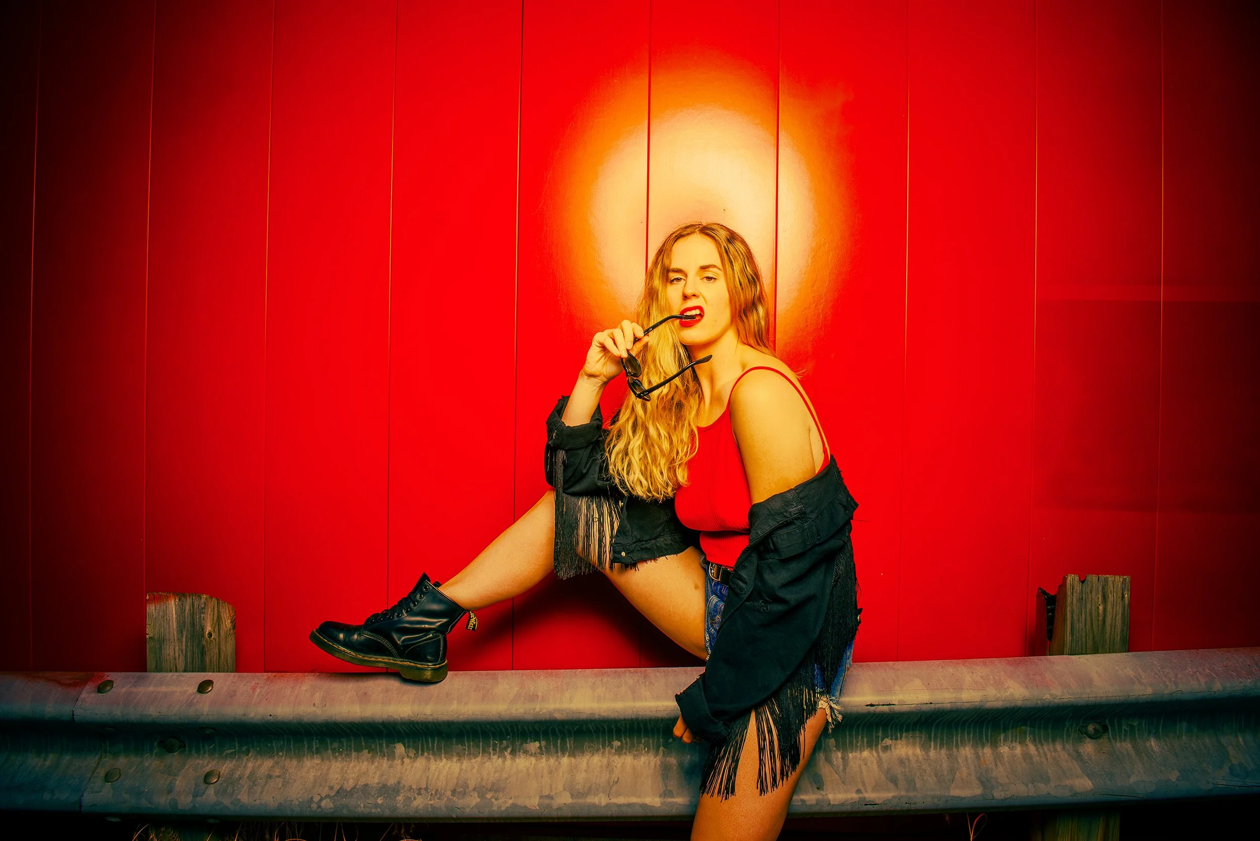Woman with long blonde curly hair posing in front of a red wooden wall, holding sunglasses, wearing a red top, black jacket, and black boots.
