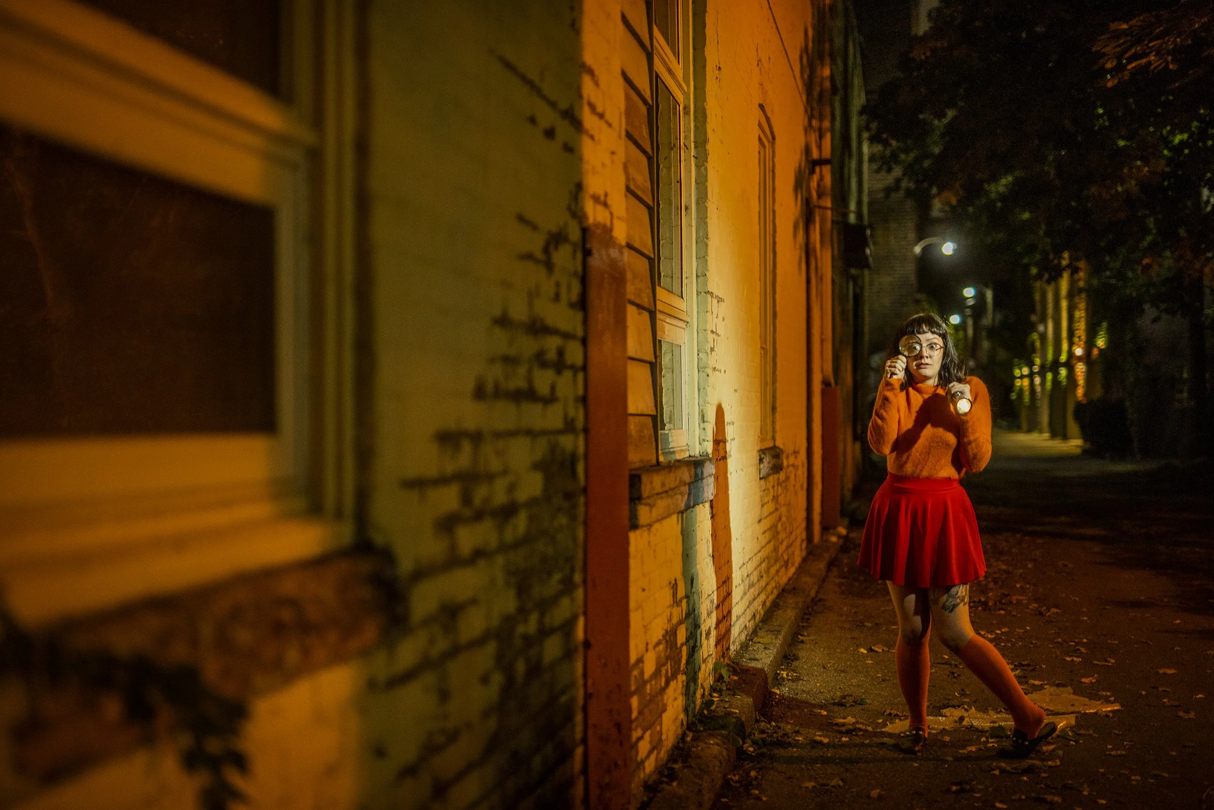 A woman in an orange sweater and red skirt wearing glasses, walking outside at night along a brick building with streetlights in the background.