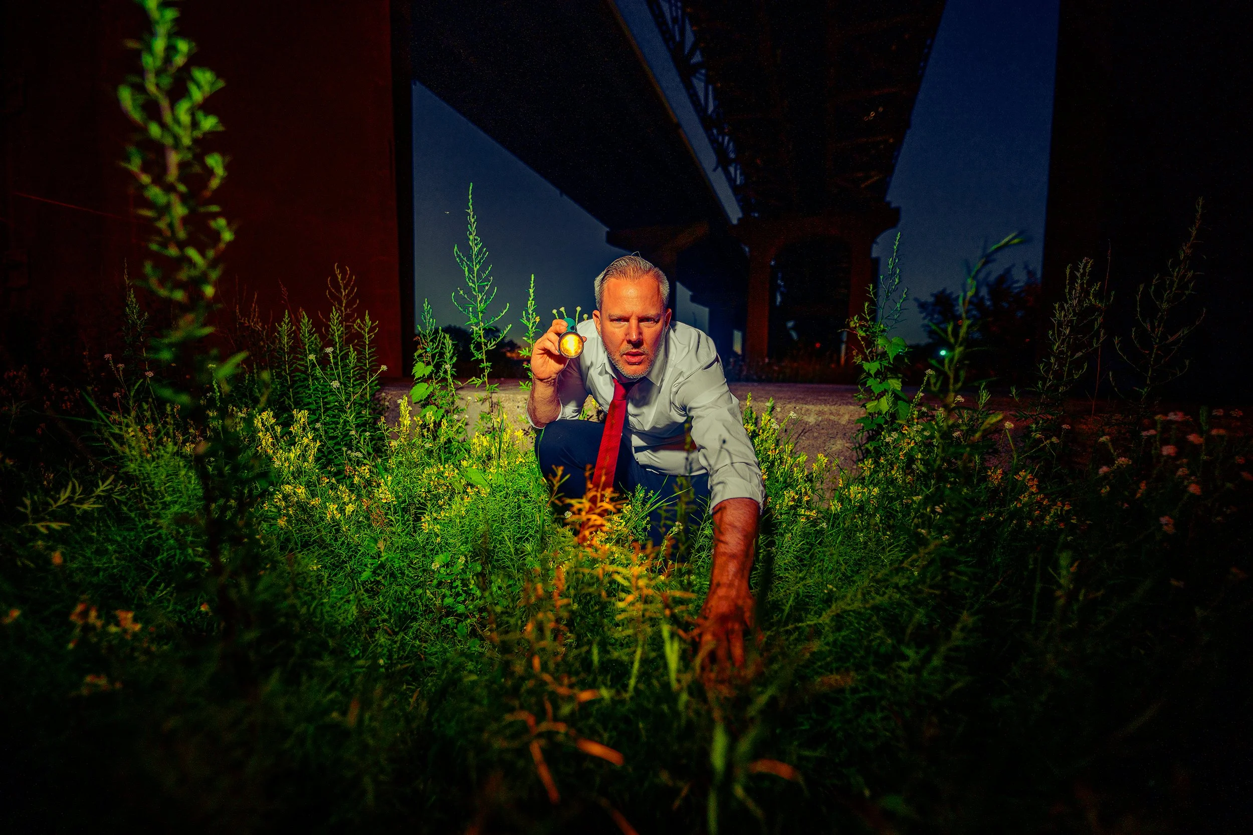A man in a white shirt and red tie crouches in a field of tall green plants under a bridge at night, holding a glowing object.