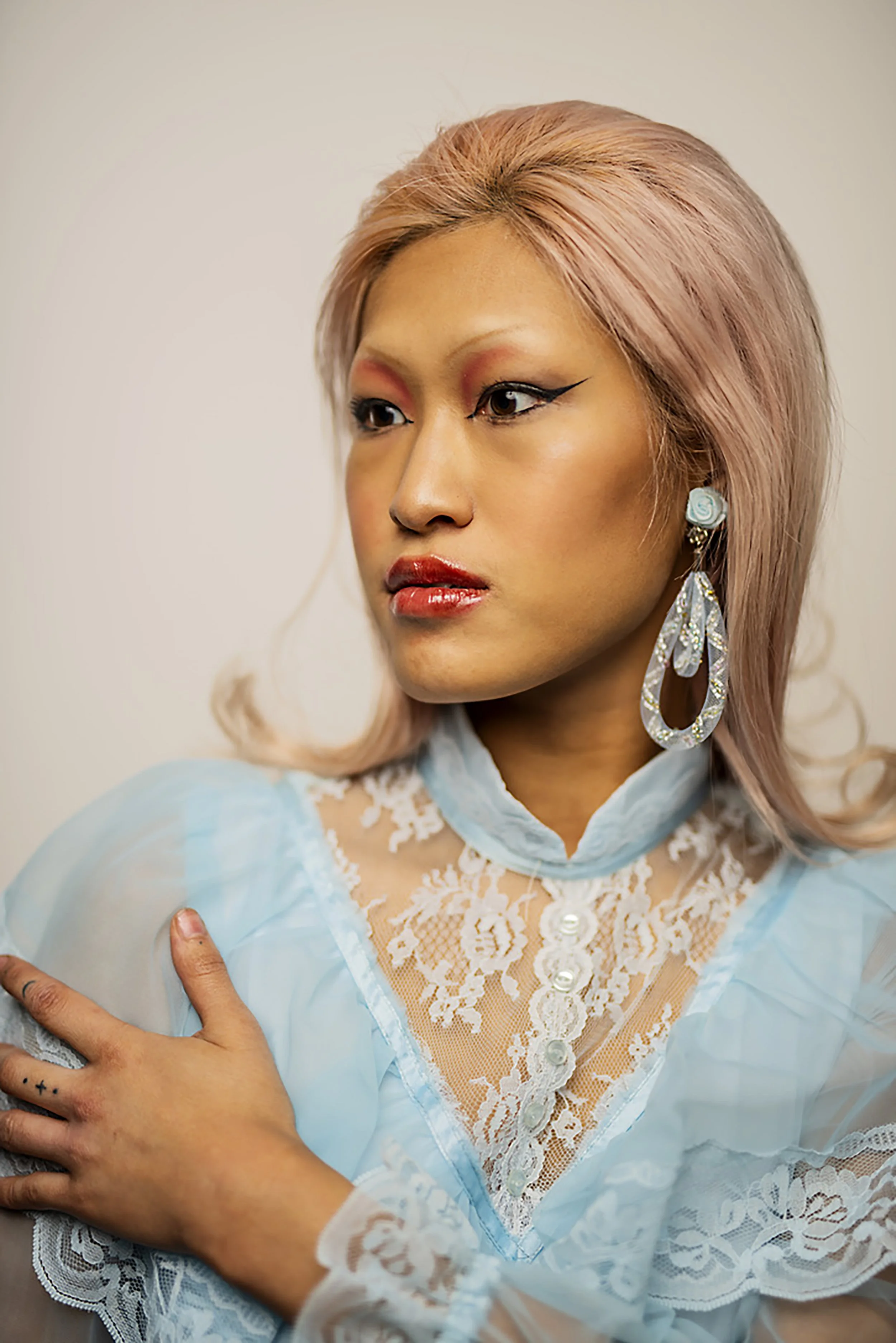 Close-up of a woman with pink hair styled with volume, wearing bold makeup with winged eyeliner and red lipstick. She has large, elegant earrings with a blue rose detail and a white lace and blue sheer top, posing with her hand on her chest against a