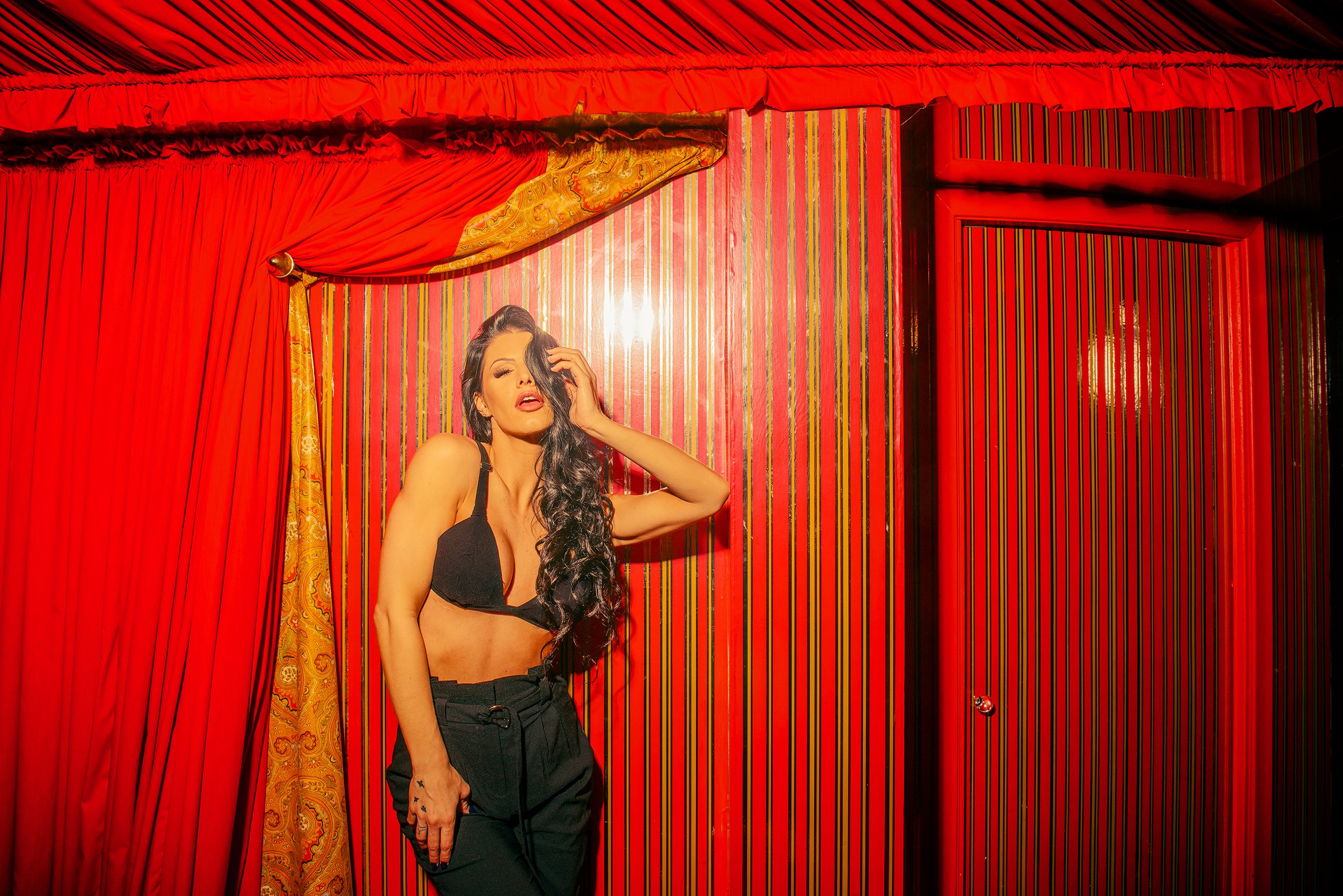 A woman with long dark hair, wearing a black bra and black pants, posing against a vibrant red background with curtains and striped walls.