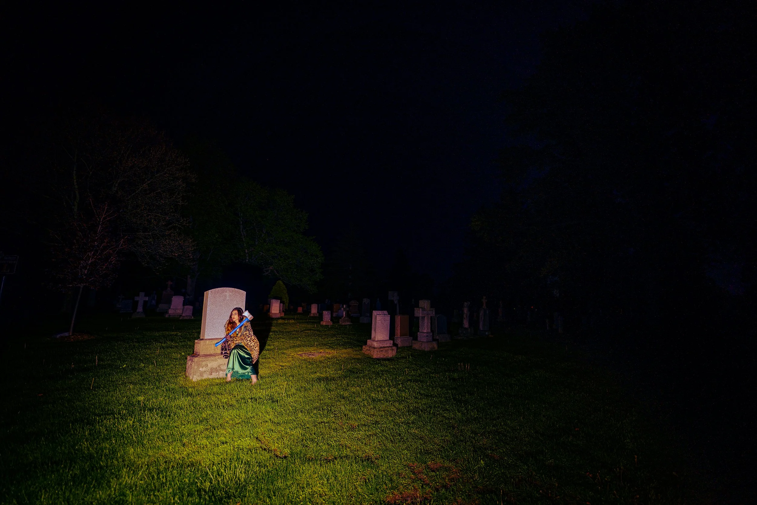 A young woman in a green skirt and leopard print top sitting on a tombstone in a graveyard at night, illuminated by a focused light, with numerous tombstones and trees in the background under a dark sky.