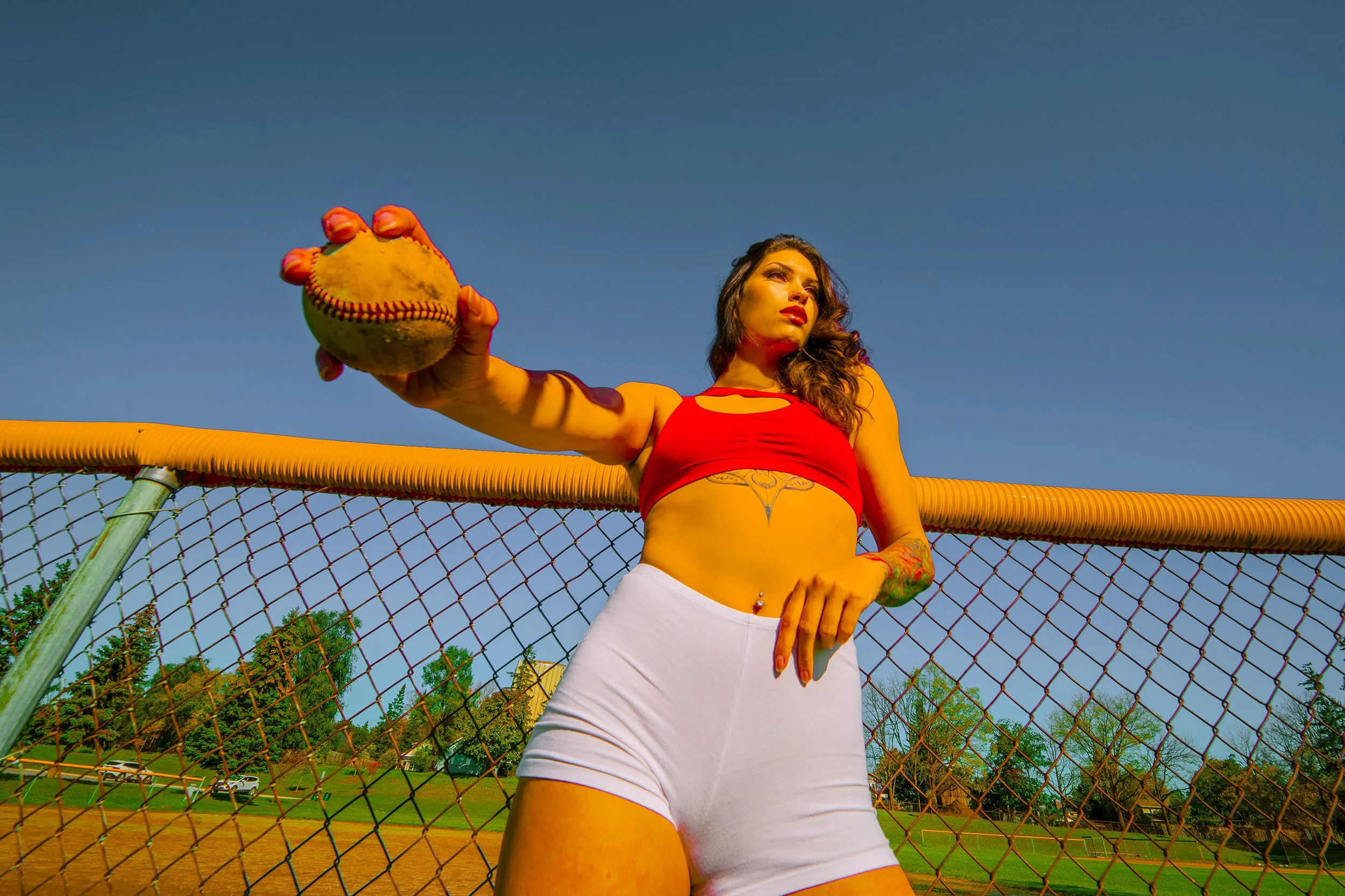A woman wearing a red sports bra and white shorts standing on a baseball field, holding a yellow softball with red stitching towards the camera, with a clear blue sky and trees in the background.