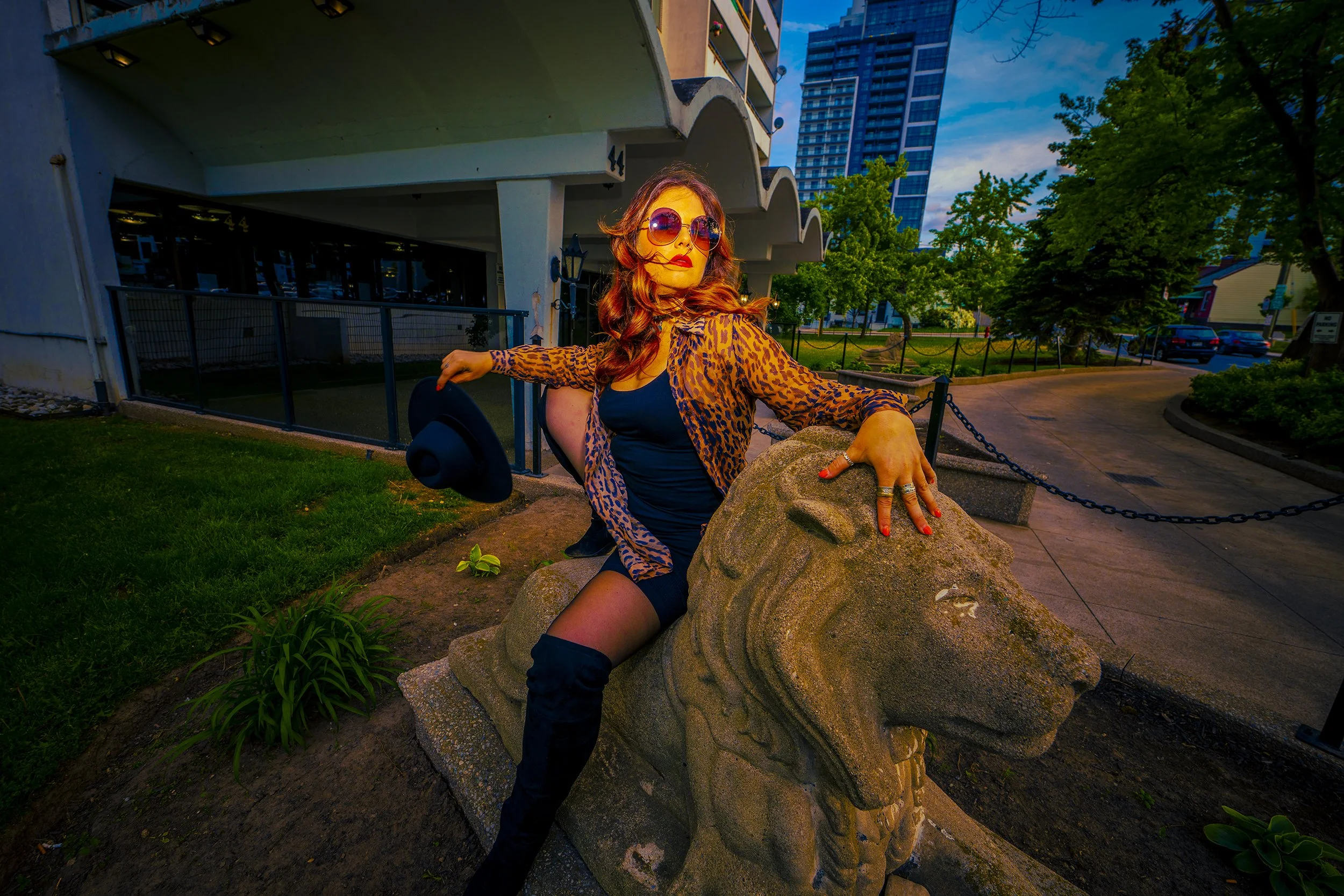 A woman with long wavy hair wearing large sunglasses and a leopard print jacket posing outdoors near a stone sculpture of a lion's face, with green trees, buildings, and a blue sky in the background.
