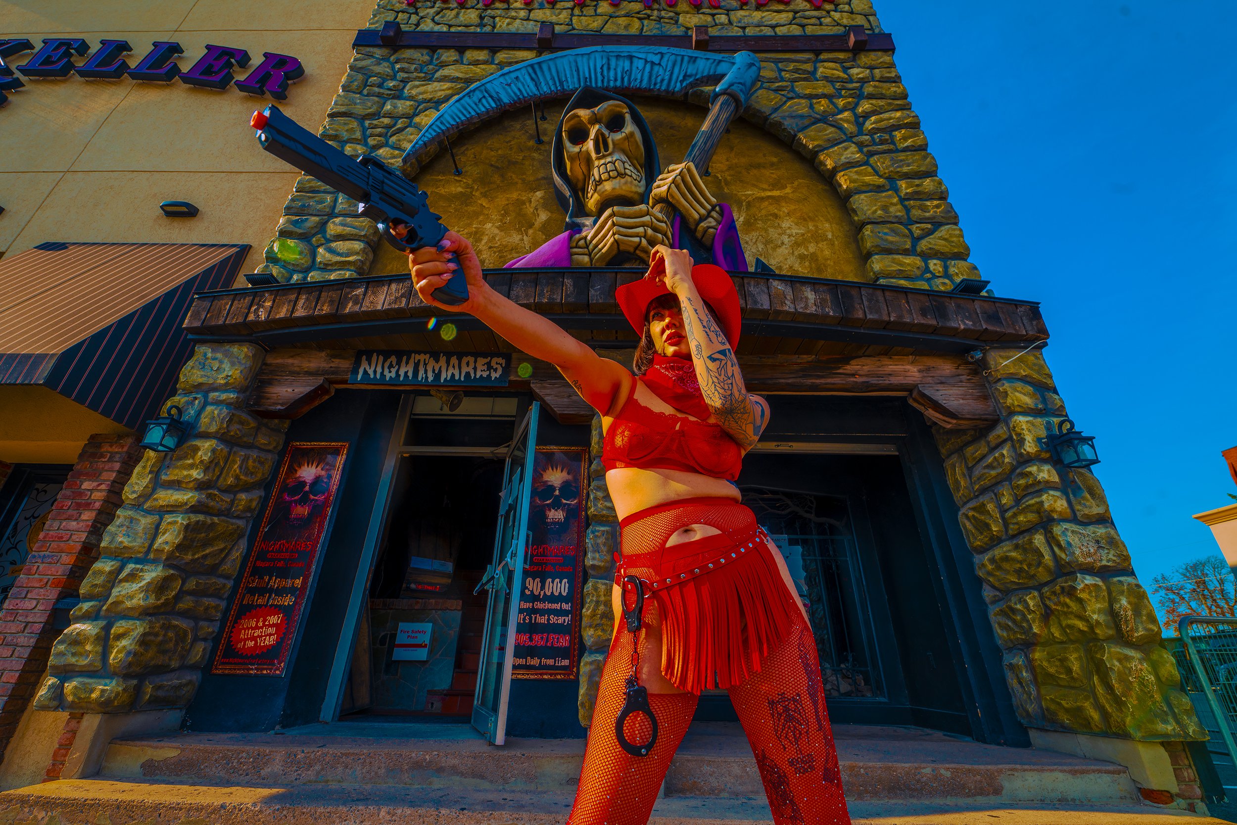 Woman dressed in red, wearing fishnet stockings and a wide-brimmed hat, stands outside a haunted house attraction named 'Nightmares,' holding a toy gun raised in the air, with a large skeleton Grim Reaper figure behind the building.