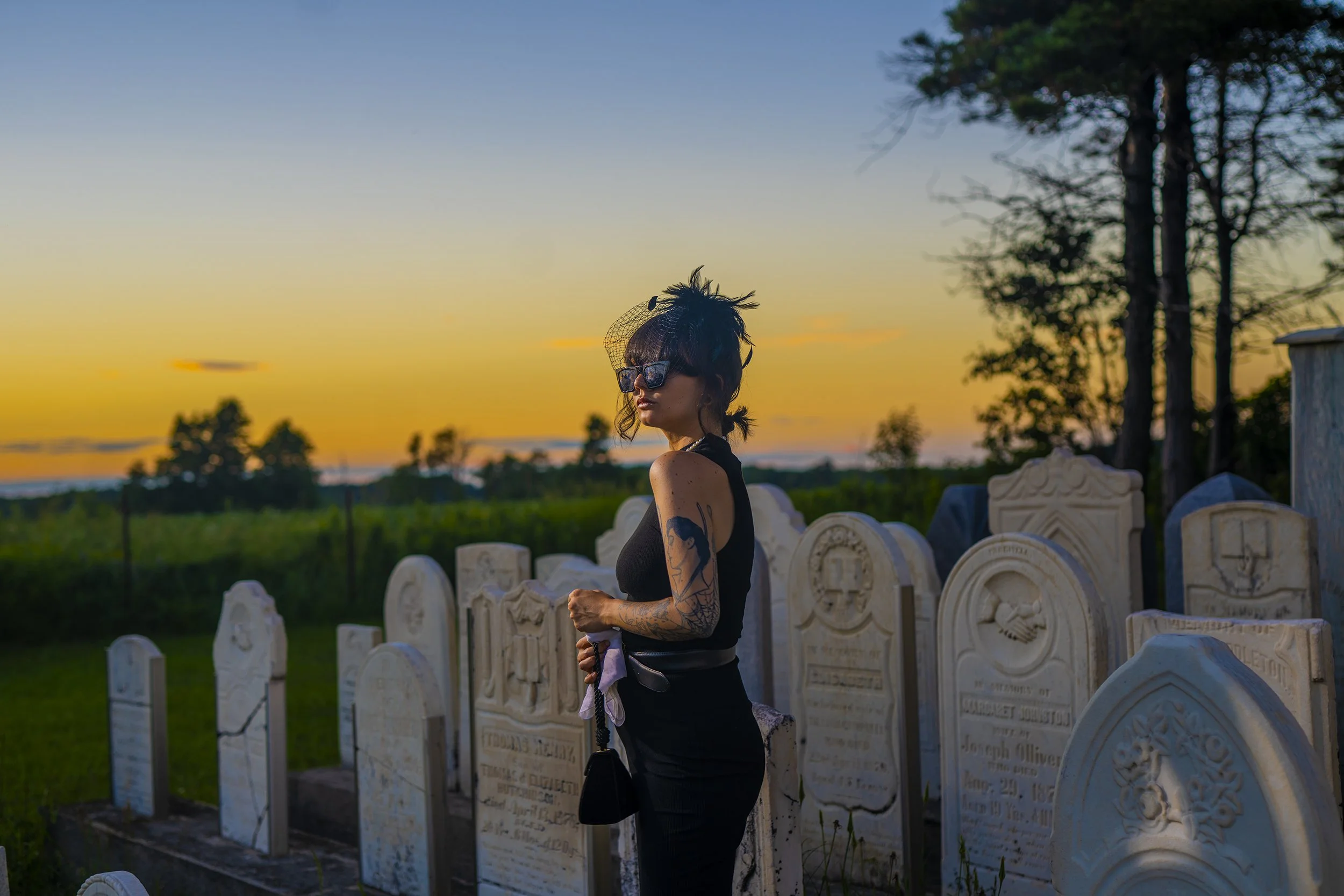 A woman with tattoos on her arm and glasses standing in a cemetery at sunset, surrounded by tombstones and trees.