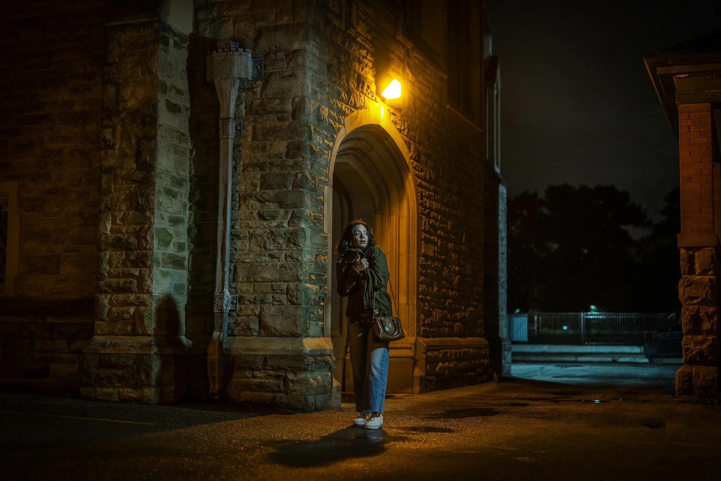 A woman standing under a yellow streetlight outside a brick building at night, with her arms crossed and looking to the side.