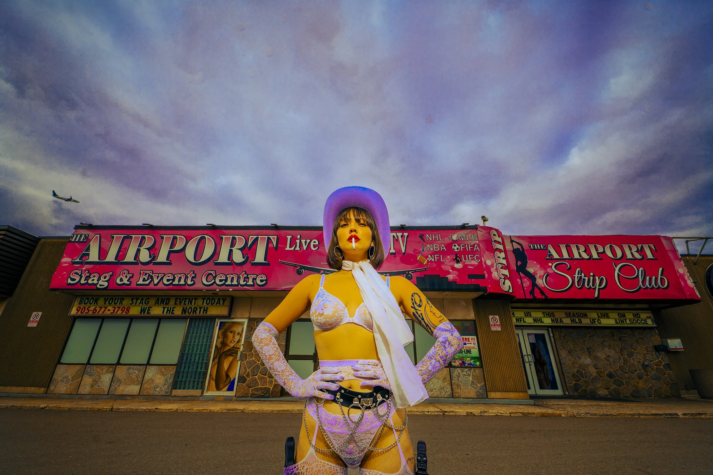 A person wearing a purple cowboy hat, white lace lingerie and gloves, with tattoos, standing in front of a pink sign for the Airport Strip Club and Stag & Event Centre, under a cloudy sky with an airplane flying overhead.
