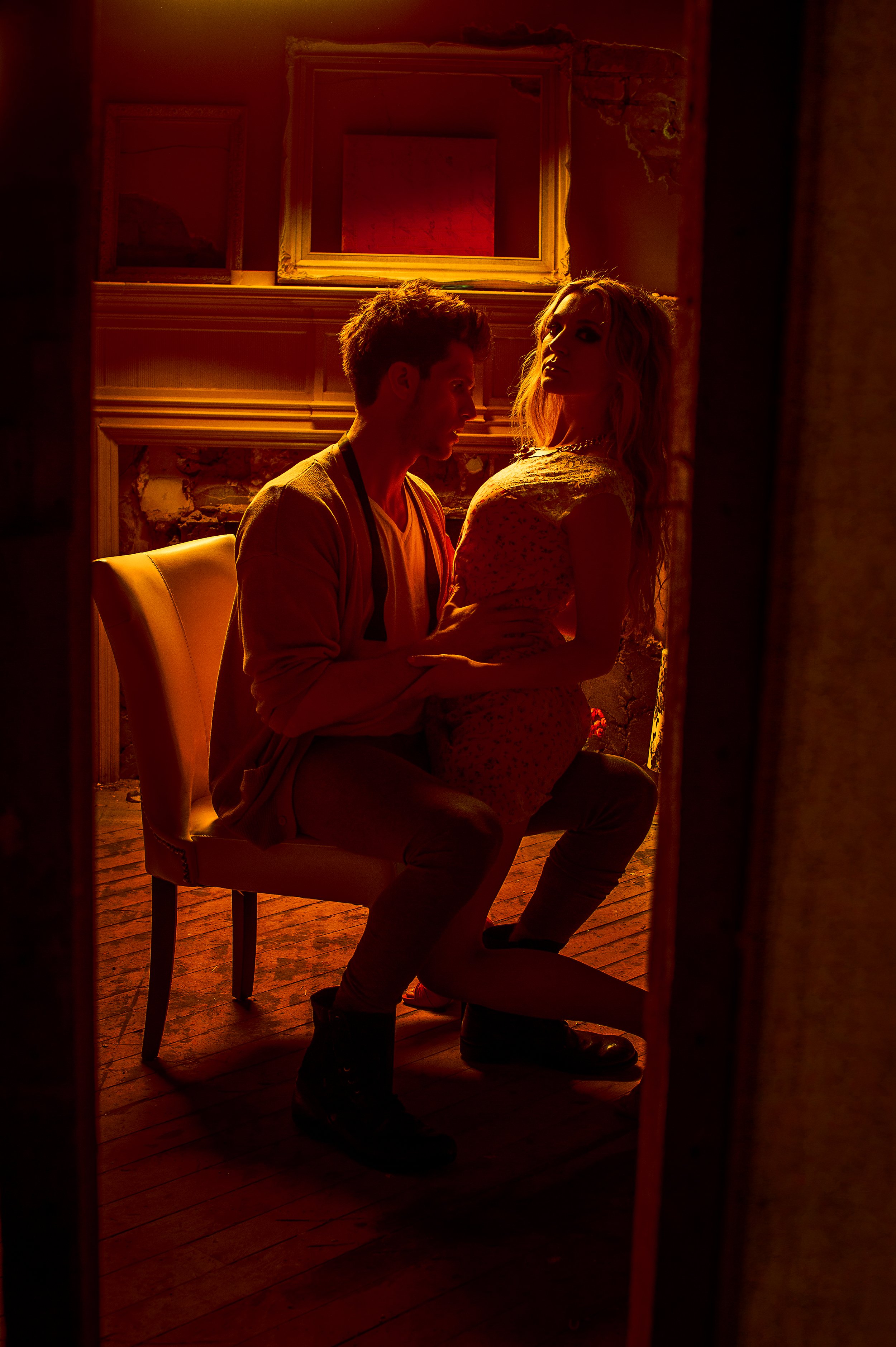 A man and woman are in a dimly lit room with red lighting, sitting close to each other. The man is seated on a chair, and the woman is kneeling in front of him, with her hand on his chest. The room has exposed brick walls and ornate picture frames on
