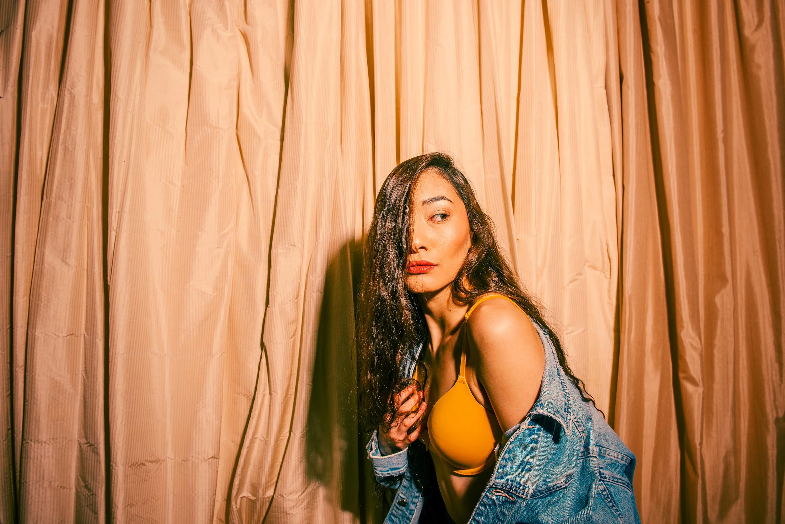 A woman with long dark hair wearing a yellow top and denim jacket sits in front of beige curtains.