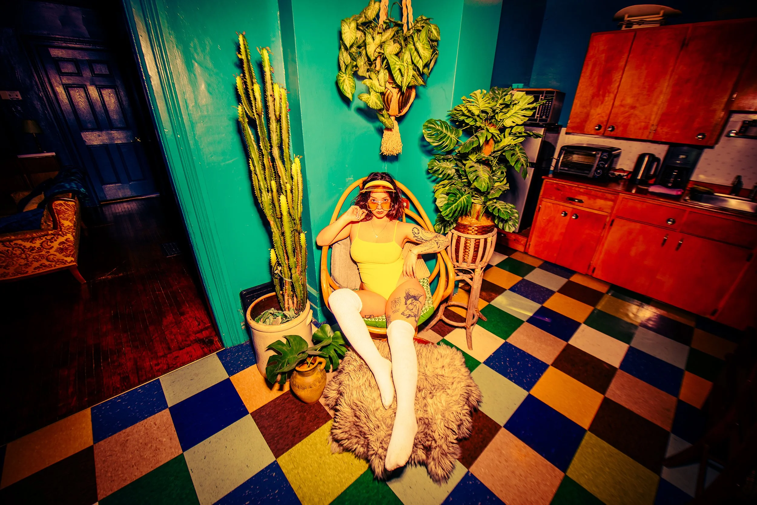 A woman with curly dark hair wearing a yellow top and white socks, sitting on a fuzzy rug on a colorful checkered floor, surrounded by tall potted plants, with a teal wall, kitchen cabinets, and various household items visible in the background.