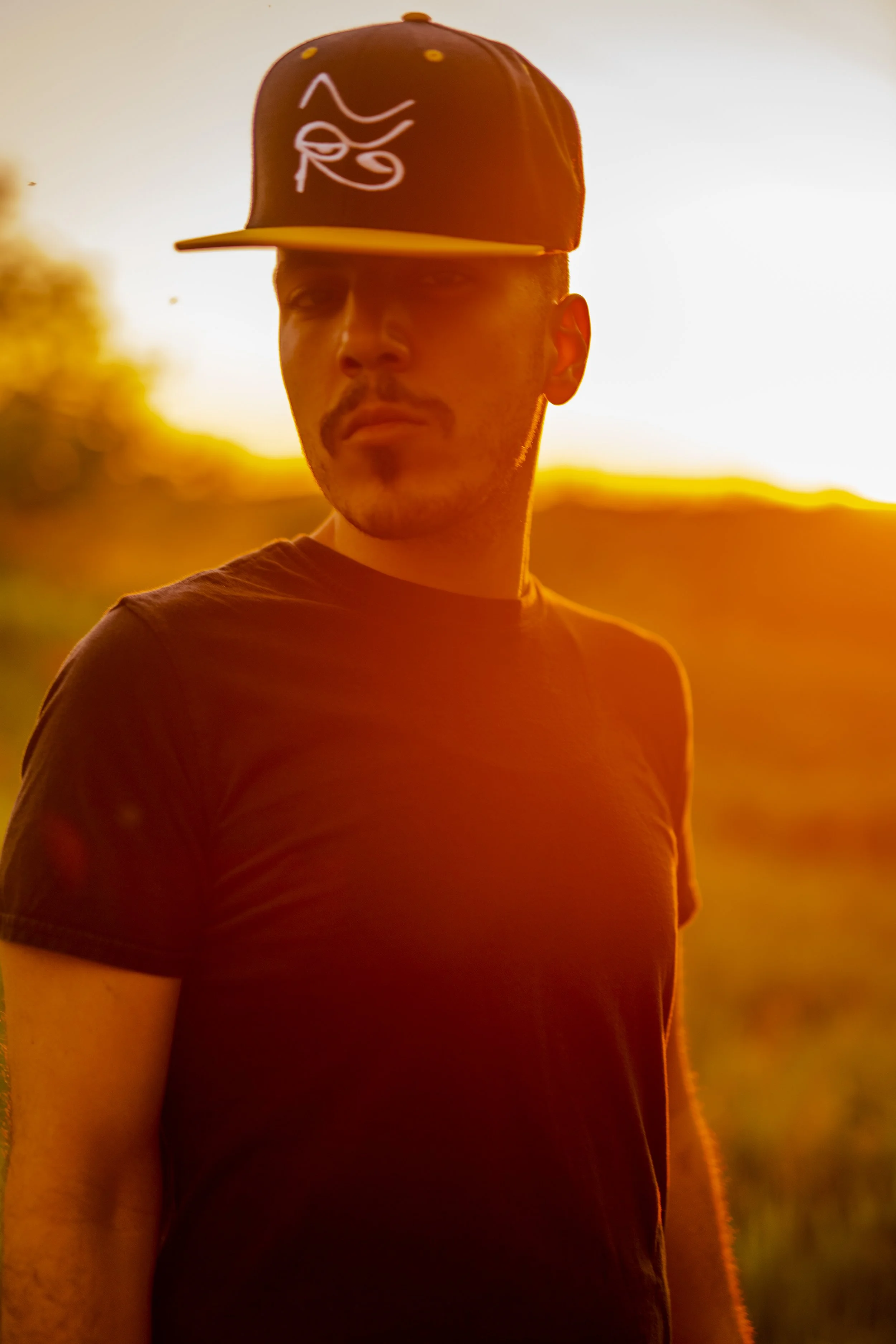 A man with facial hair wearing a black T-shirt and a black baseball cap with a white logo, standing outdoors during sunset with a warm, orange glow in the background.
