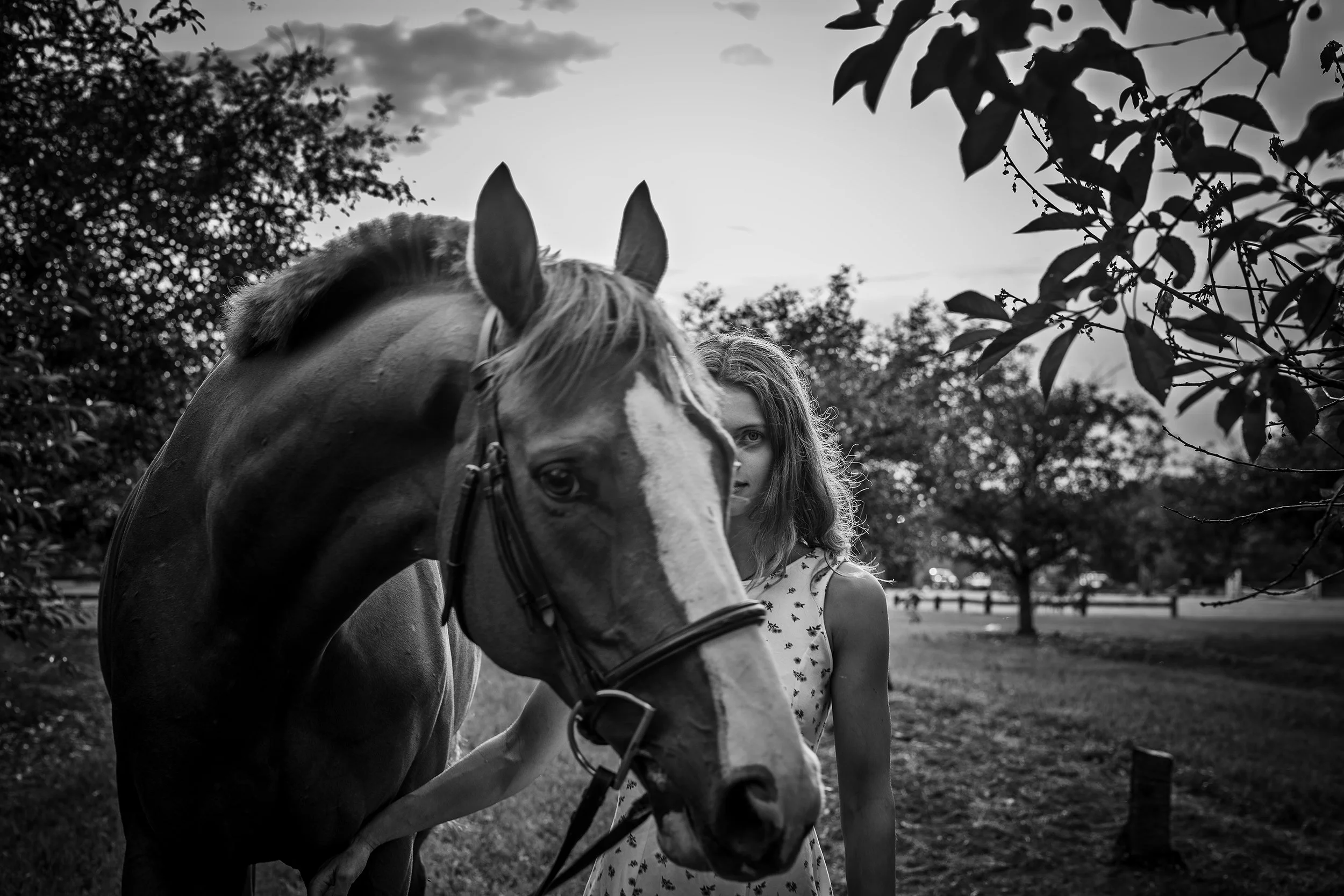 A black-and-white photograph of a young woman walking alongside a horse in a park. The woman has long, wavy hair and is wearing a patterned dress. The horse, which is close to the camera, has a light-colored coat with a darker mane, and is wearing a 