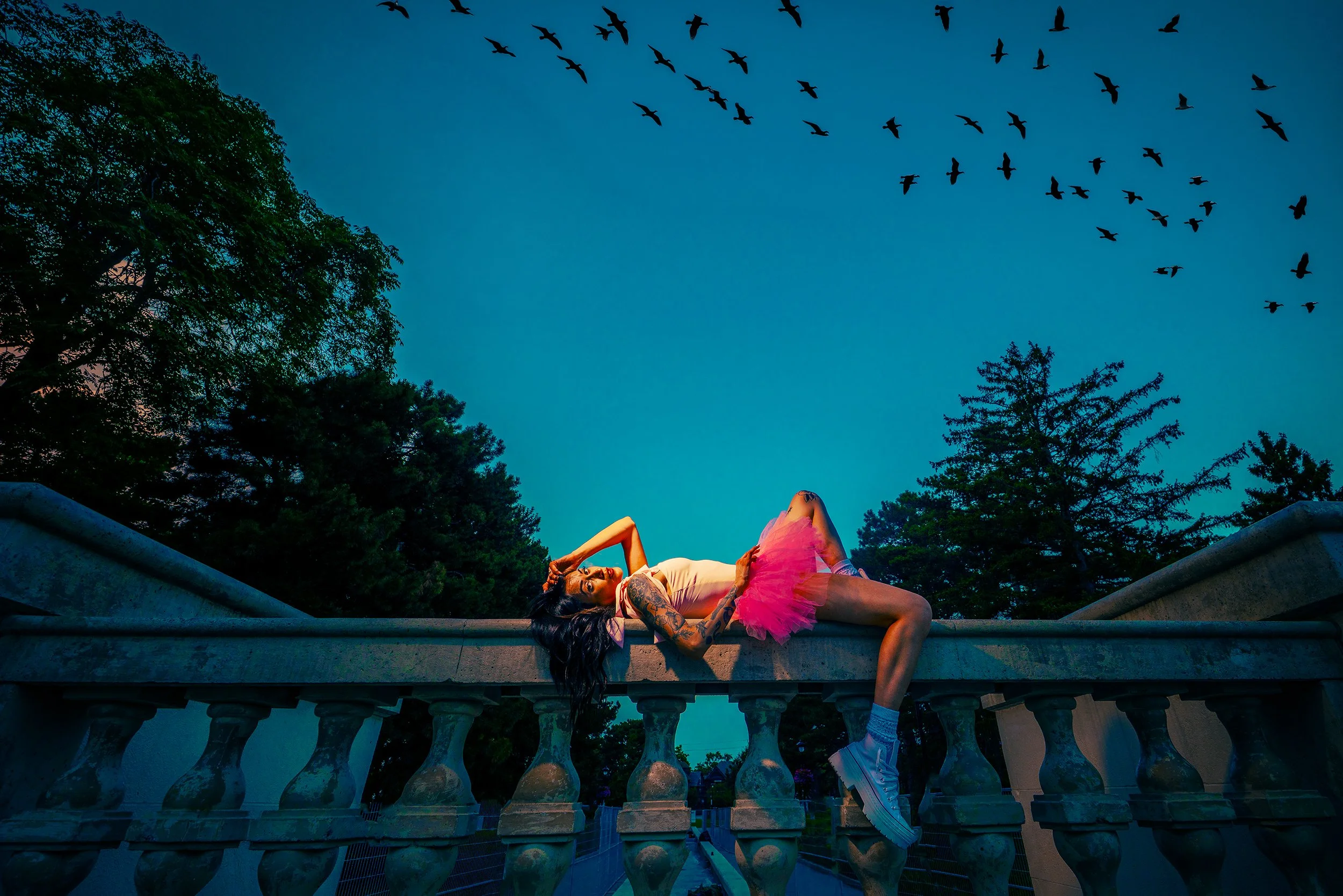 A woman lying on a stone balustrade at dusk, wearing a pink tutu, white t-shirt, and white sneakers, with dark hair and tattoos on her arm, in a park surrounded by green trees and flying birds above.