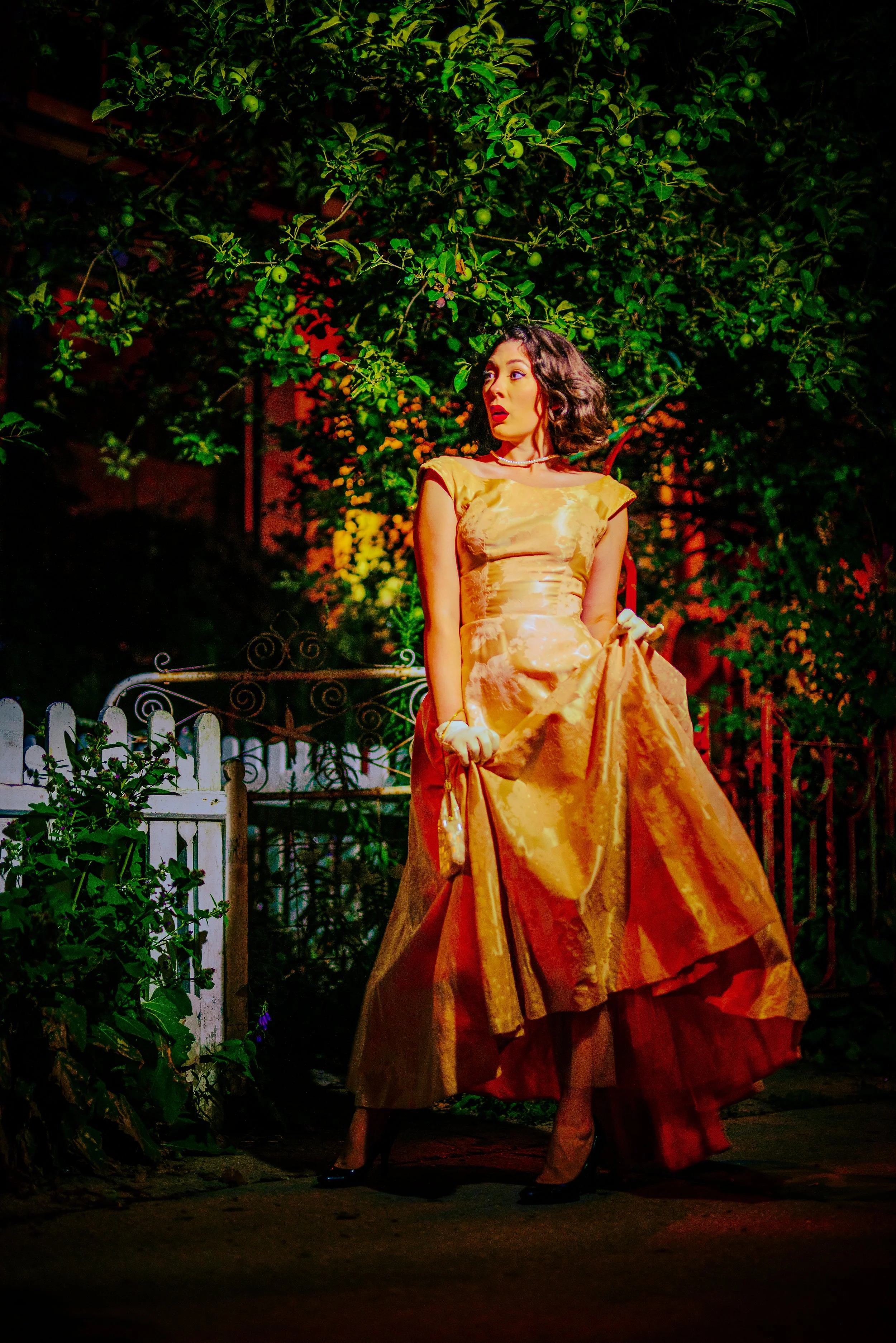 A woman dressed in a long, elegant, golden yellow gown standing outdoors at night, with a lush green tree in the background. She has dark wavy hair and is wearing high heels.