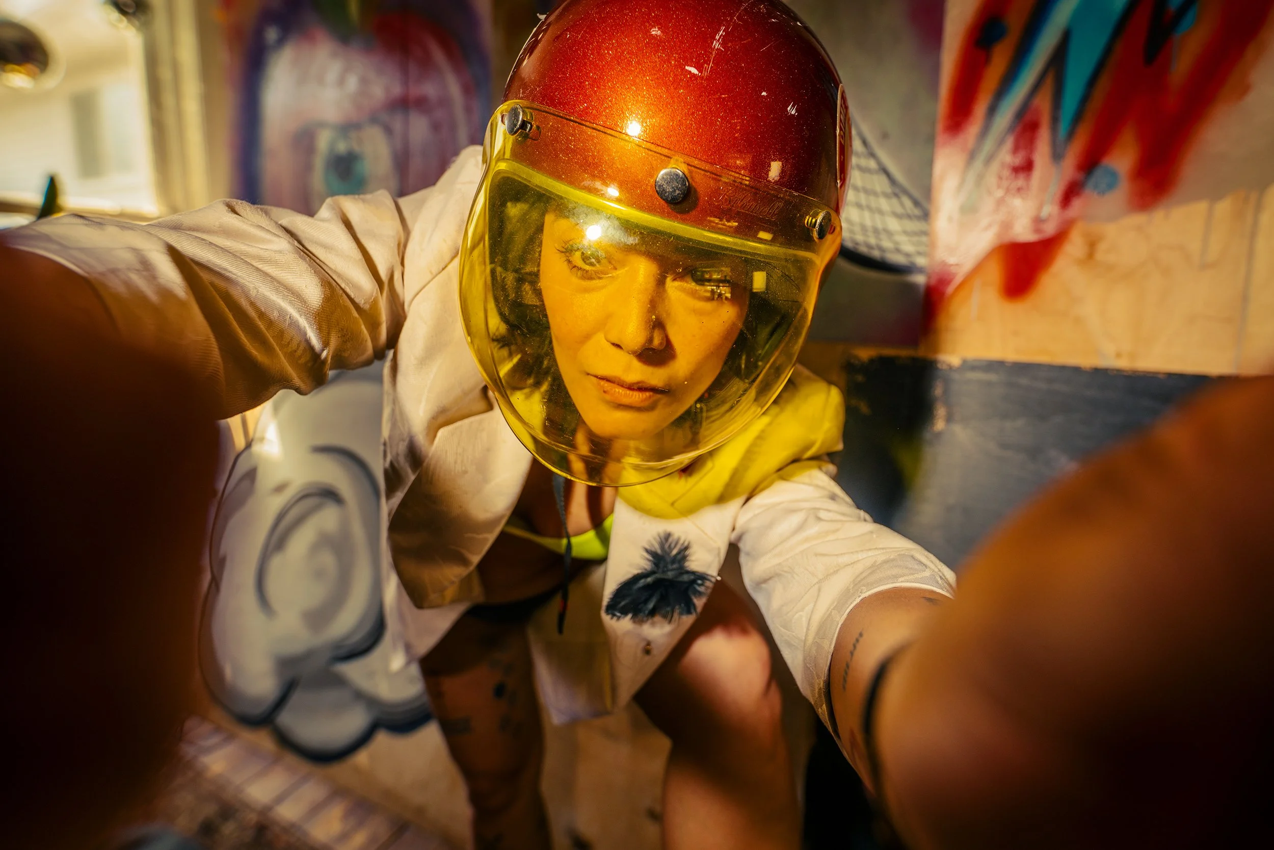 A woman wearing a shiny red helmet with a yellow face shield and a white shirt with black designs is taking a selfie from a low angle. The background features colorful graffiti art.