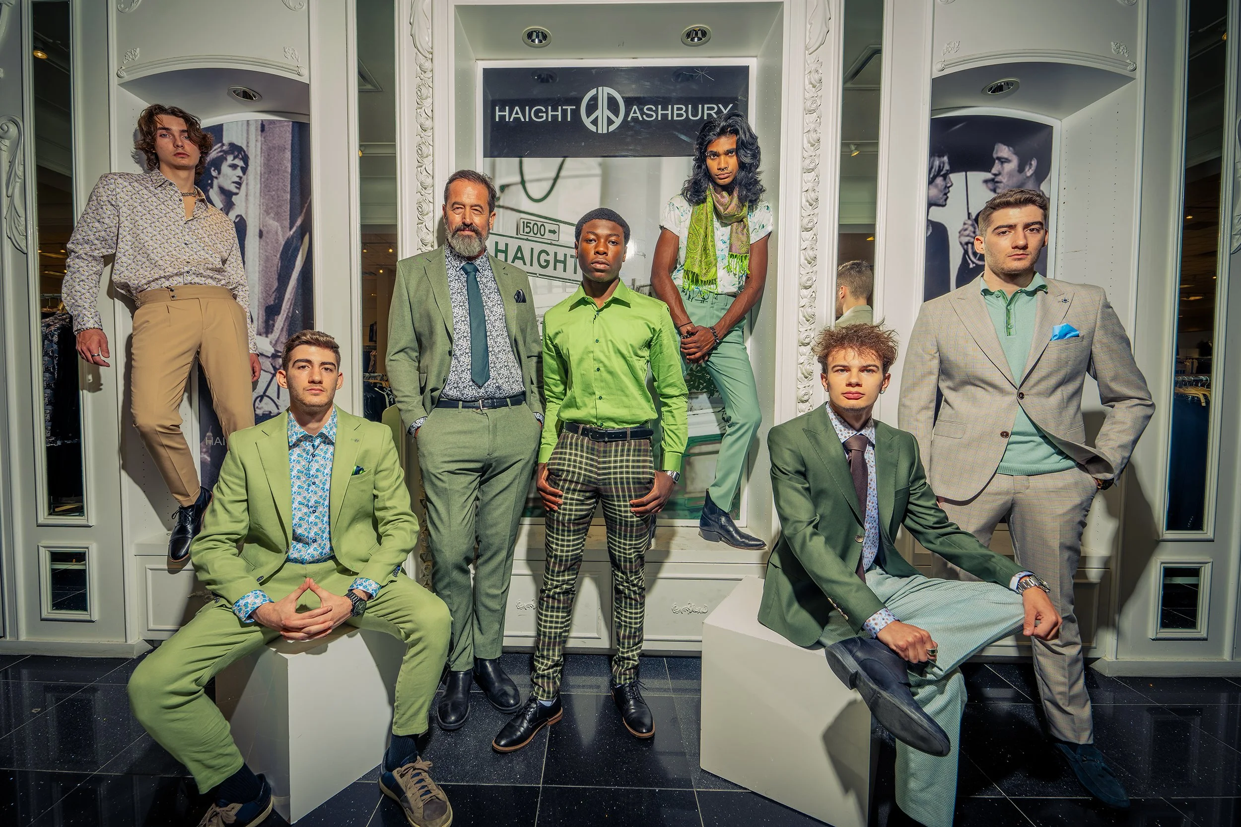 A group of seven fashionably dressed men posing in front of a storefront with a sign that reads 'Haight Ashbury' and black-and-white portraits of couples on the walls.