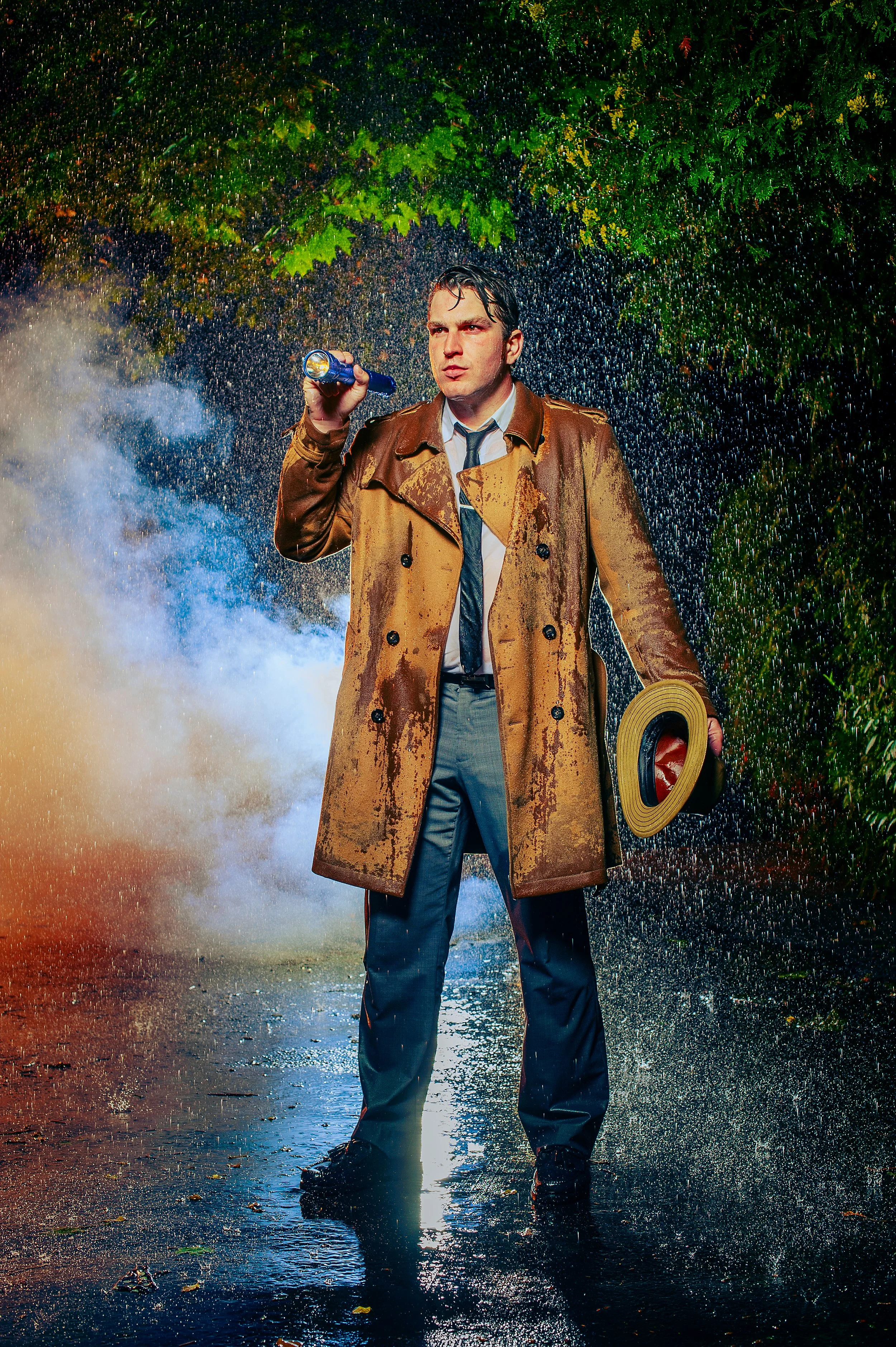 A man standing in the rain at night, wearing a wet trench coat and holding a flashlight and a hat, with a background of green leaves and wet pavement.