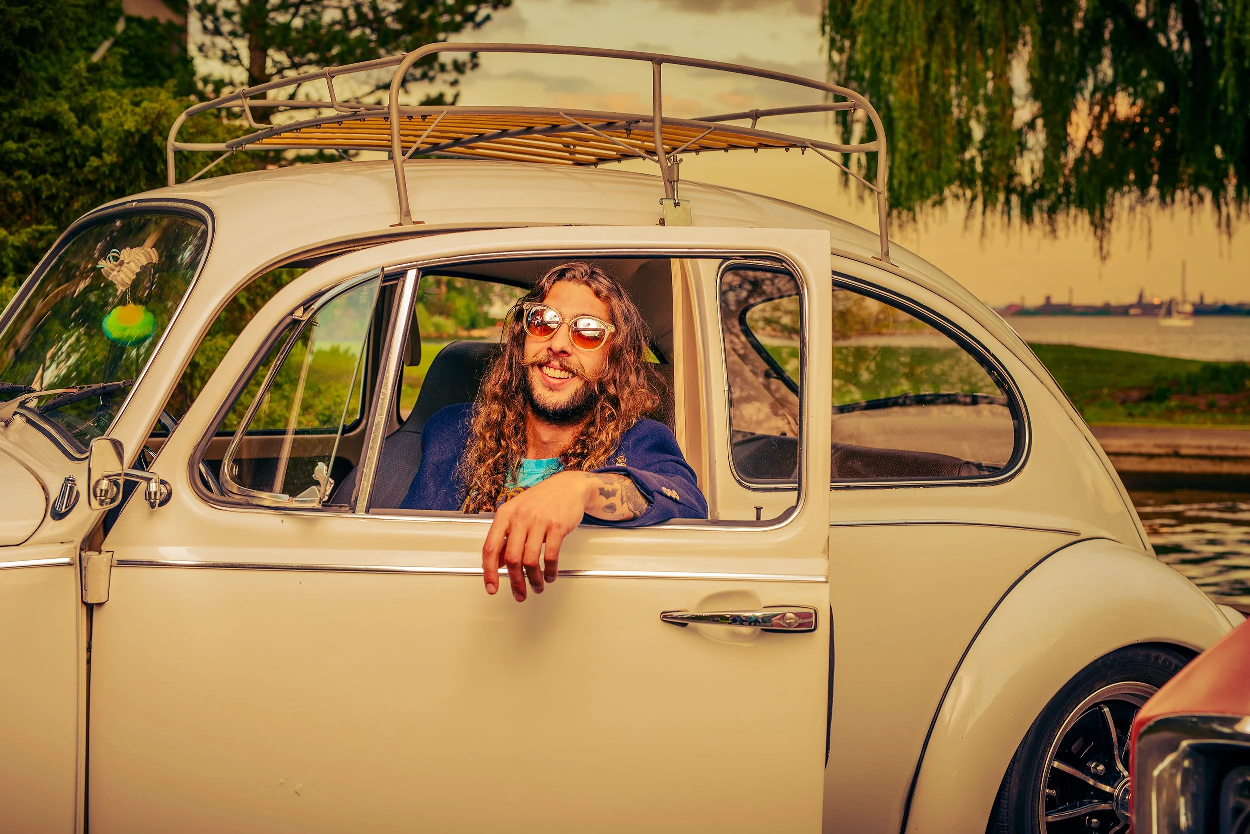 A man with curly hair and wearing sunglasses is sitting in the driver's seat of a vintage cream-colored car with a roof rack. The car is parked outdoors near a body of water with trees and a sunset sky in the background.