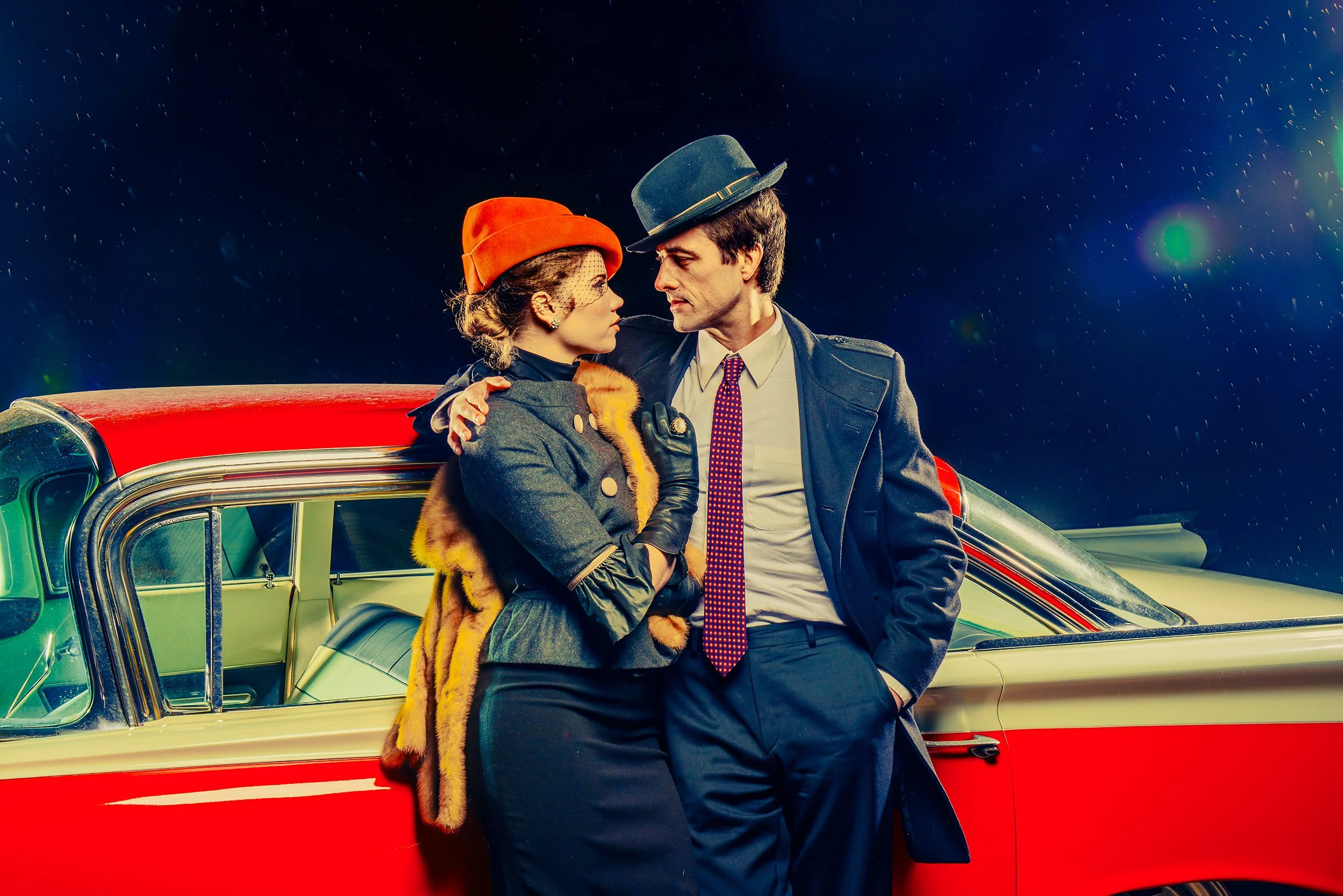 A man and a woman stand close together in front of a vintage red and cream car under a starry night sky. The woman wears a red hat, a gray coat with fur details, and black gloves, while the man wears a gray hat, a dark coat, a white shirt, and a red 