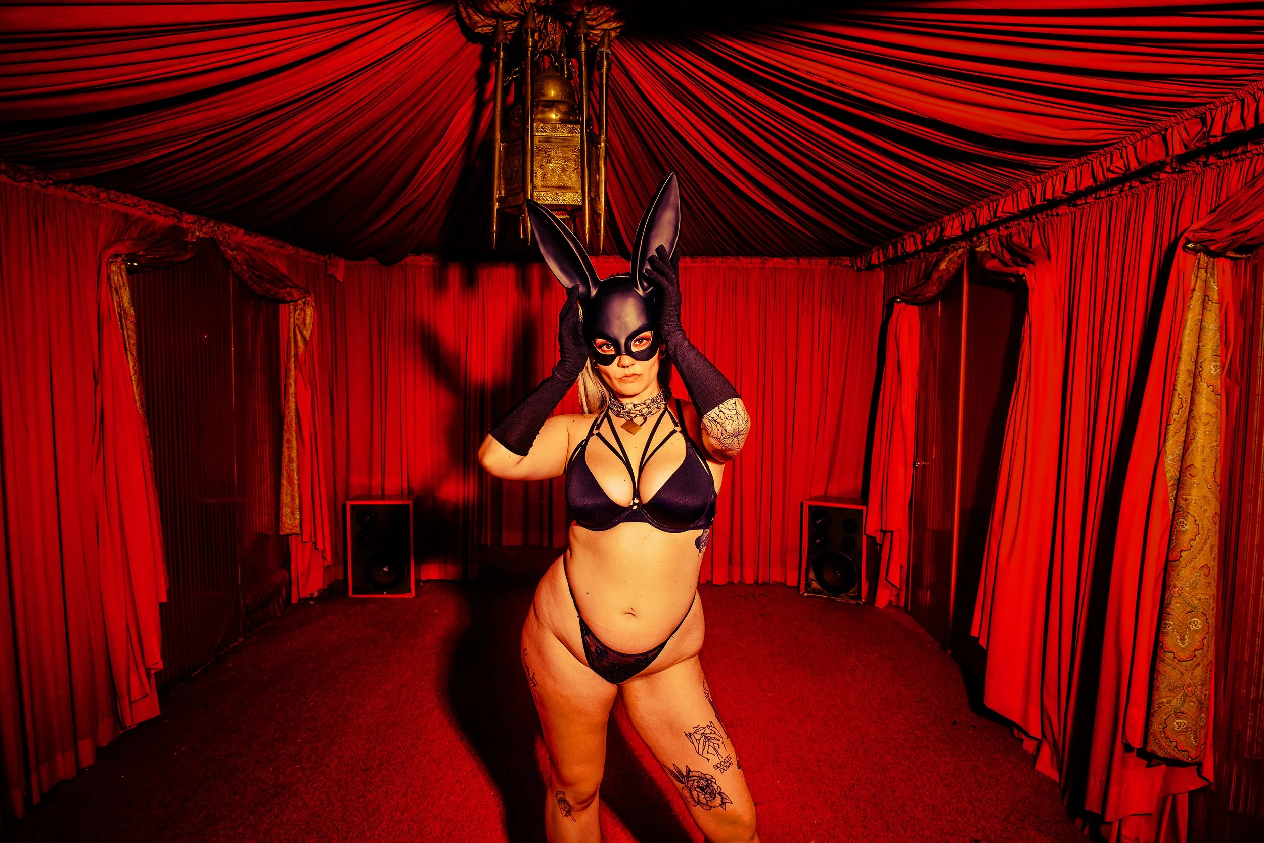 A woman wearing a black bunny mask, black lingerie, and gloves standing in a red curtained room, with tattoos on her arm and leg.