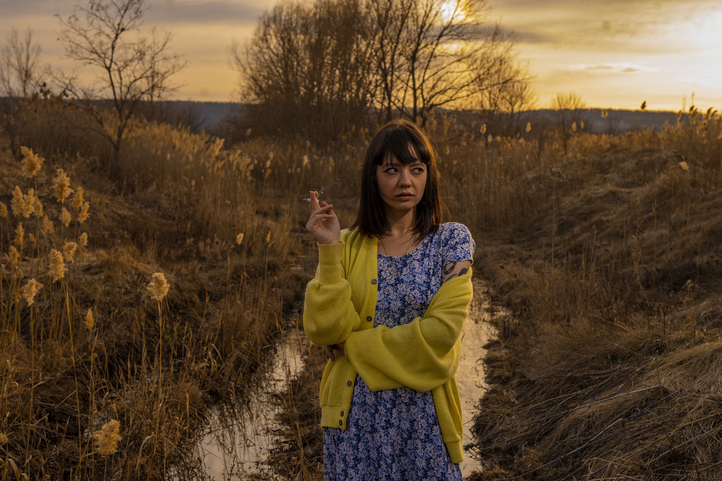 A woman standing outdoors during sunset, holding a cigarette in her right hand, wearing a yellow cardigan over a blue floral dress, with a river and autumn trees in the background.