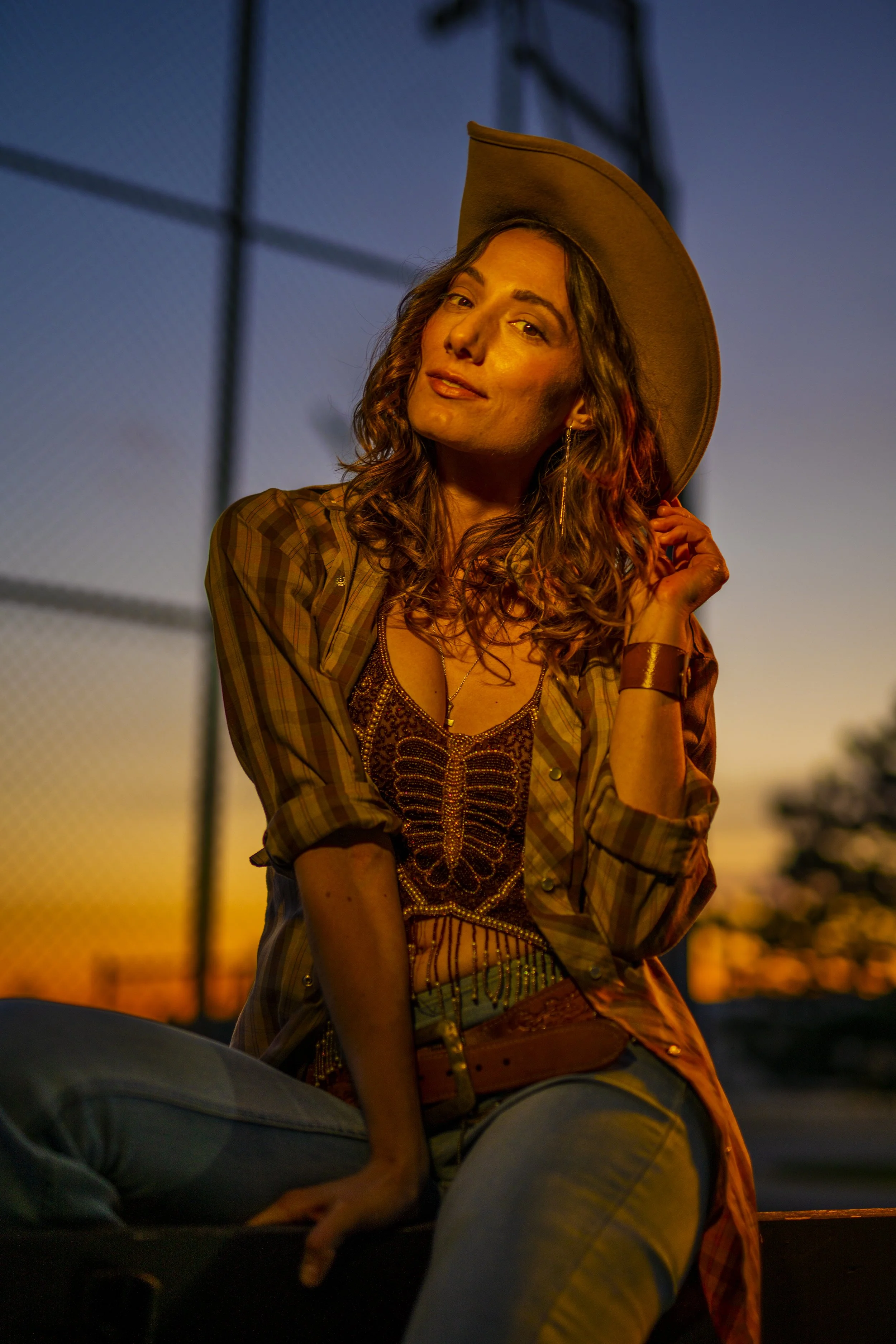 A woman with curly hair wearing a cowboy hat and casual clothing, posing outdoors at sunset near a chain-link fence.