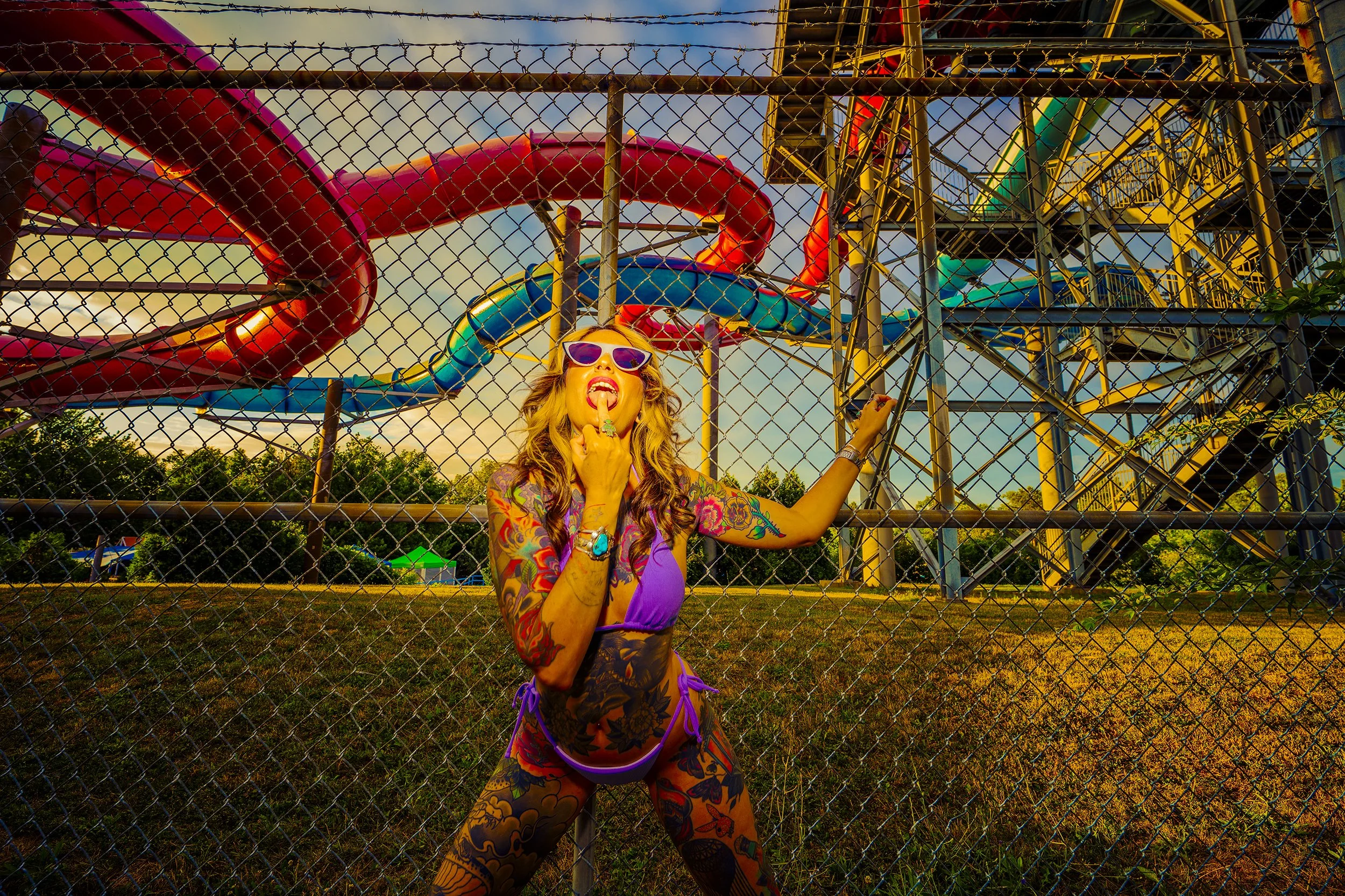 A woman with tattoos and sunglasses standing behind a chain-link fence at an amusement park, posing with her tongue out and finger on her lips, with colorful water slides and a ferris wheel in the background during sunset.