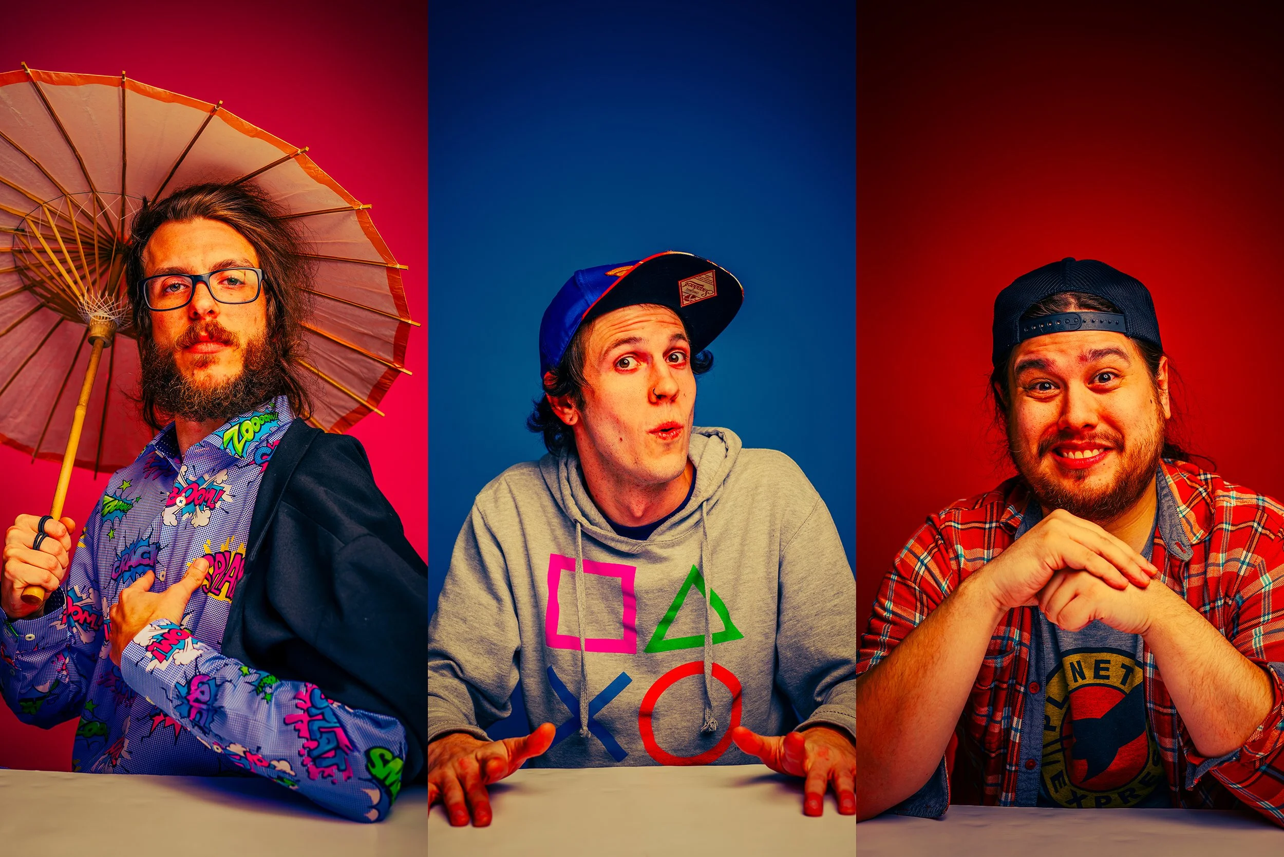 Three young men sitting at a table in front of colorful backgrounds. The man on the left holds an umbrella and wears glasses and a comic book-themed shirt. The man in the middle wears a hoodie with PlayStation symbols and a baseball cap, gesturing wi