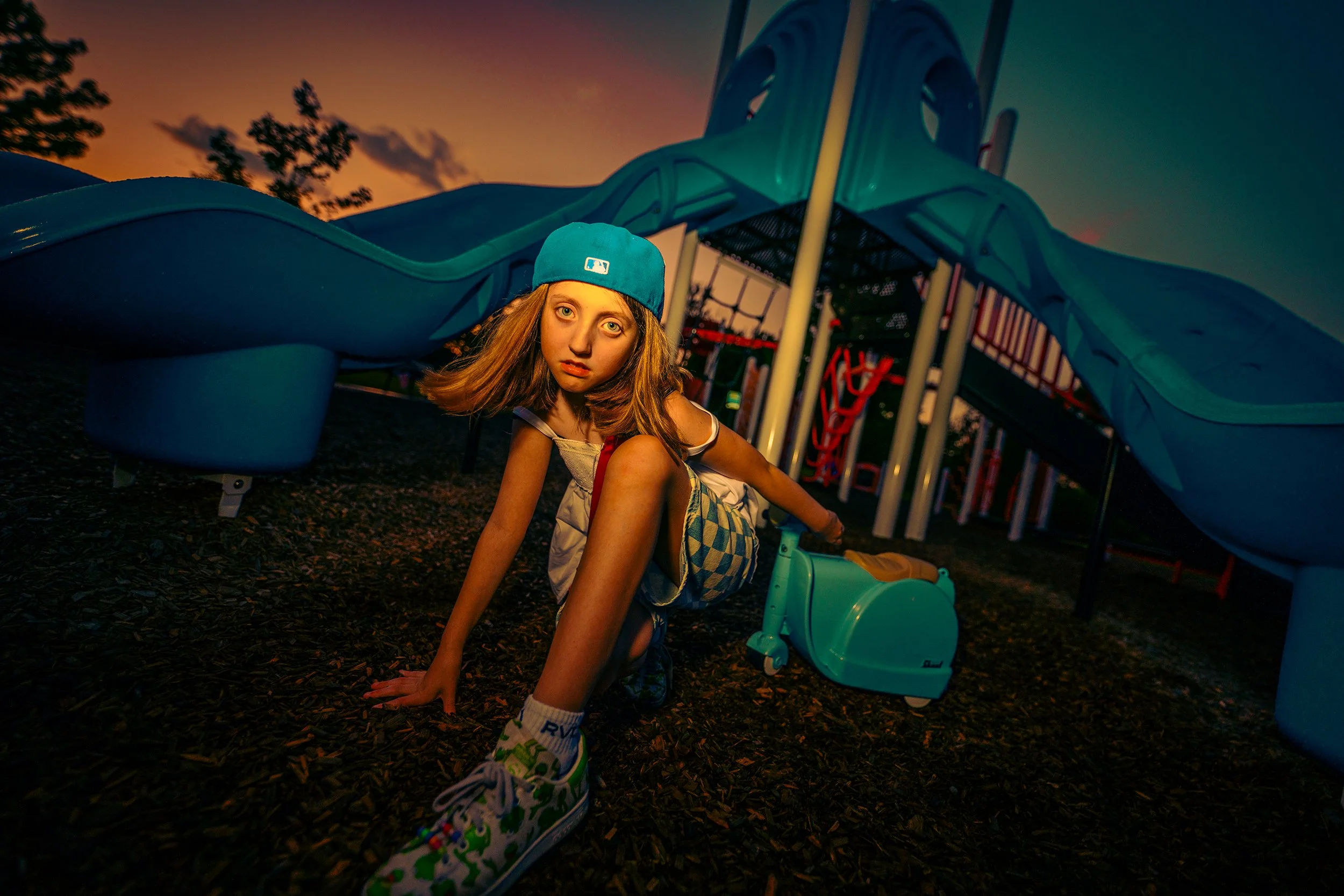A young girl crouches underneath a playground slide at sunset, looking at the camera, wearing a blue baseball cap and colorful sneakers.