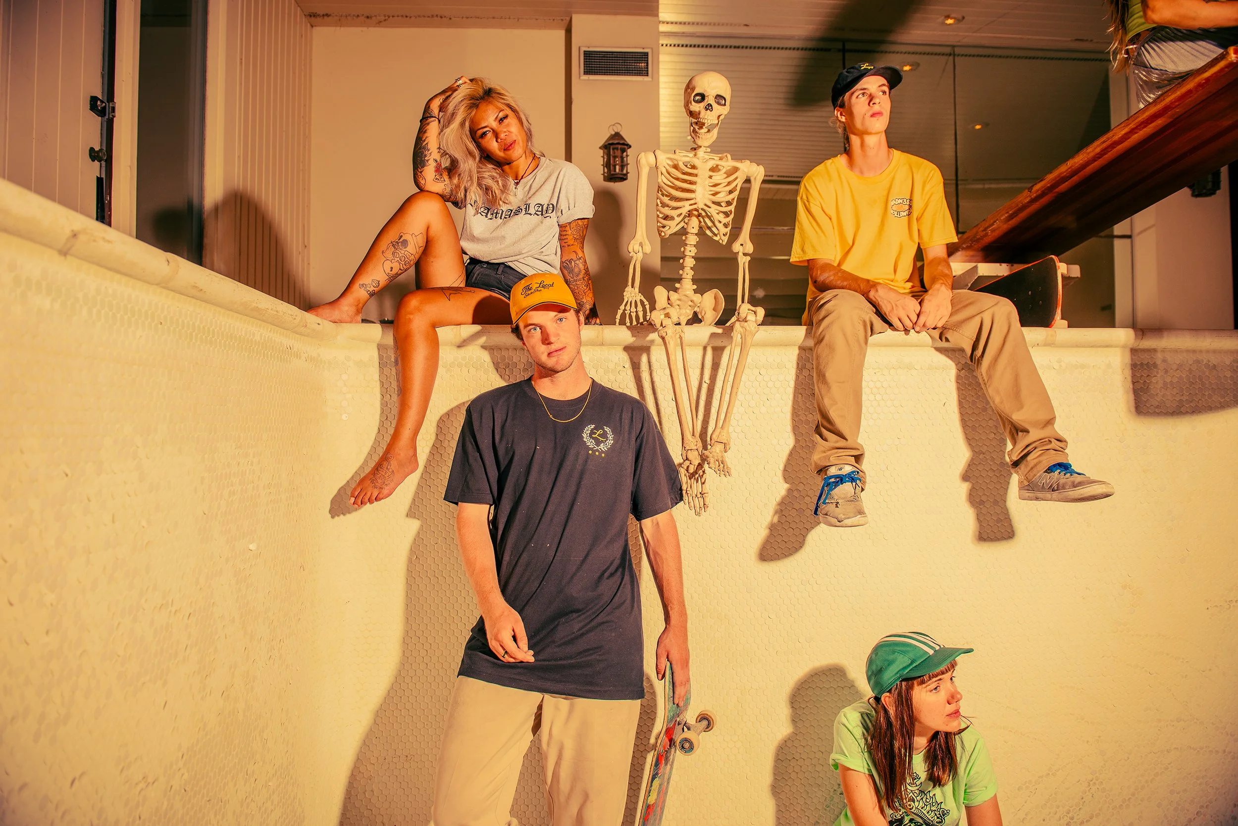 Four young people sitting and standing on a pool edge, with a human skeleton model and skateboard nearby. The woman on the left is blonde with tattoos, wearing a grey t-shirt, sitting with her leg over the pool edge. The man on the right is wearing a