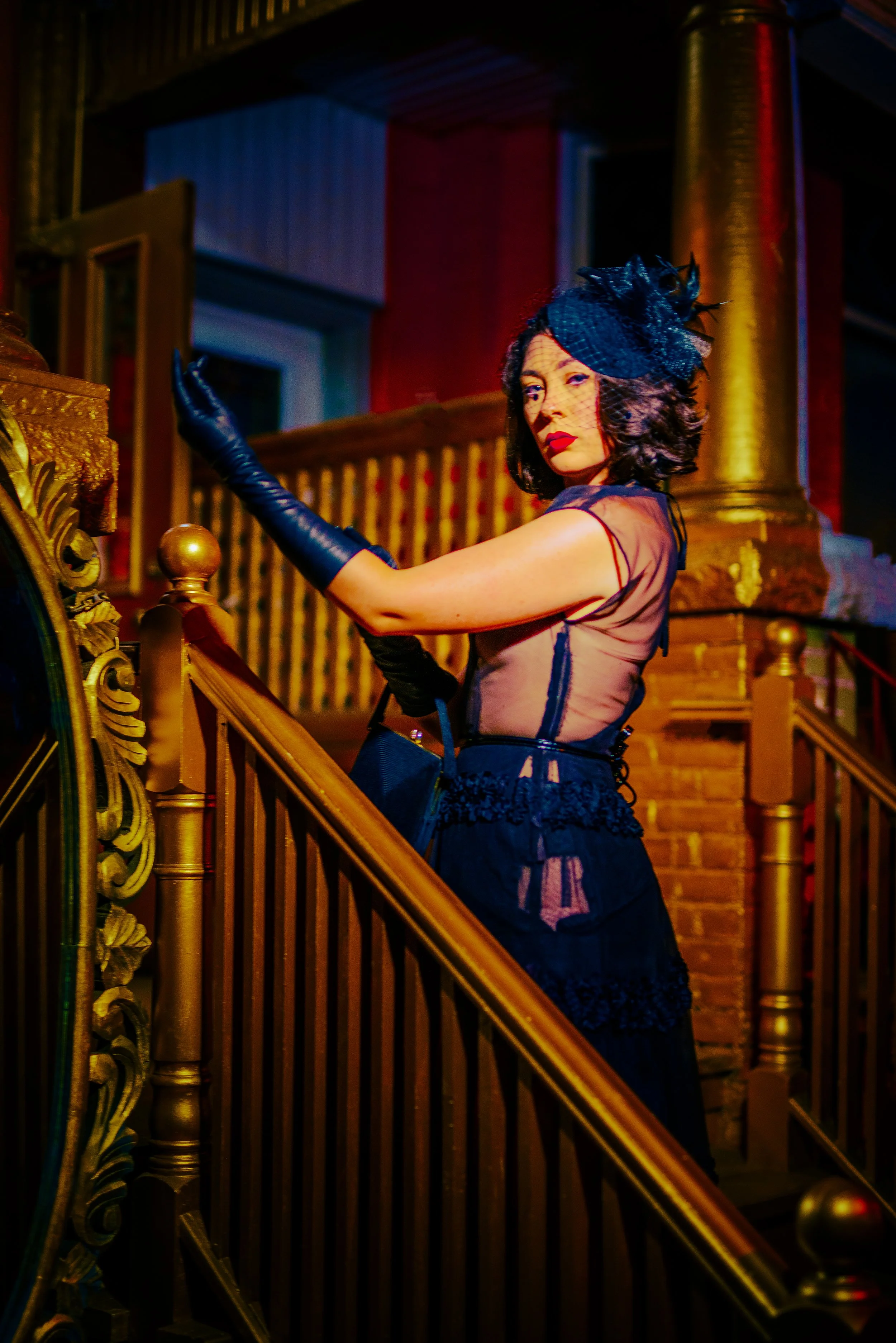 A woman with dark, curly hair wearing a black fascinator hat with netting and feathers, black gloves, and a sheer black dress with lace details, standing on a staircase with ornate gold railing, in a warmly lit, elegant interior setting.