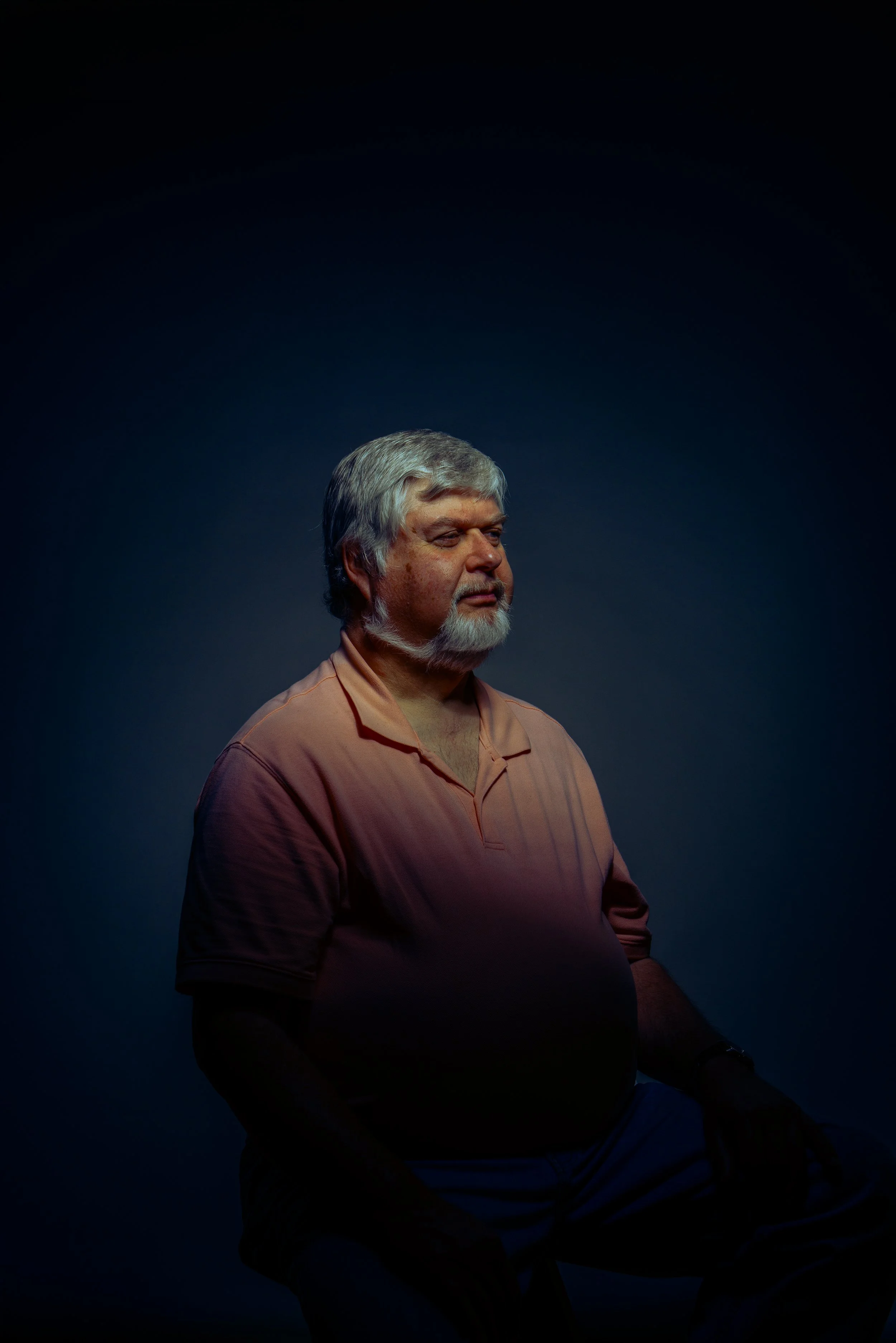 A middle-aged man with gray hair and a beard sitting in a dimly lit room, wearing a salmon-colored shirt, looking to the side with a calm expression.