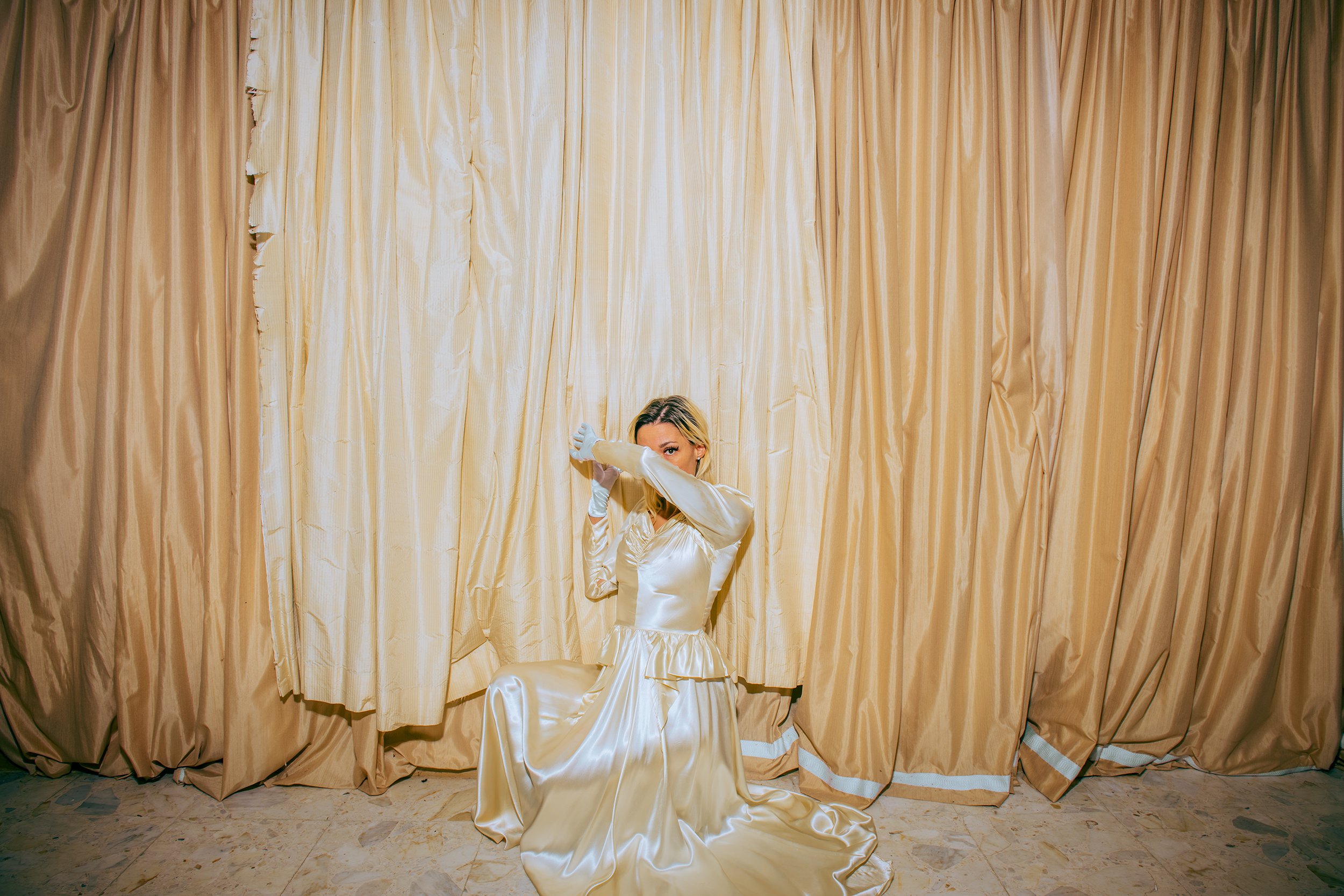 A woman in a shiny, satin, long sleeve dress is sitting on the floor in front of a large, gold curtain. She is wearing white gloves and has one arm raised to her face, partially covering it.
