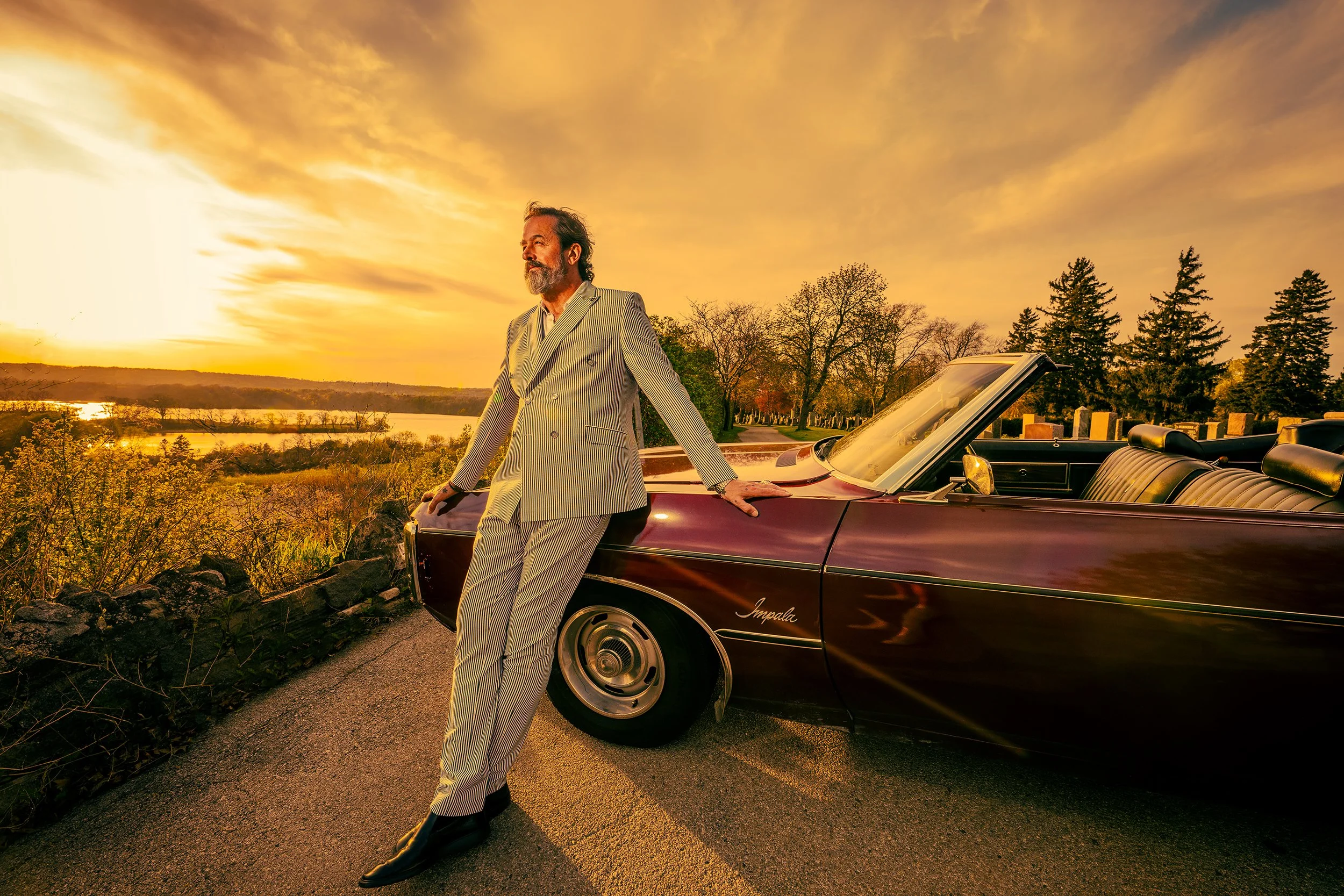 A man with a gray beard and long hair in a striped suit standing next to a vintage maroon convertible car during sunset, with a landscape of trees and a river in the background.