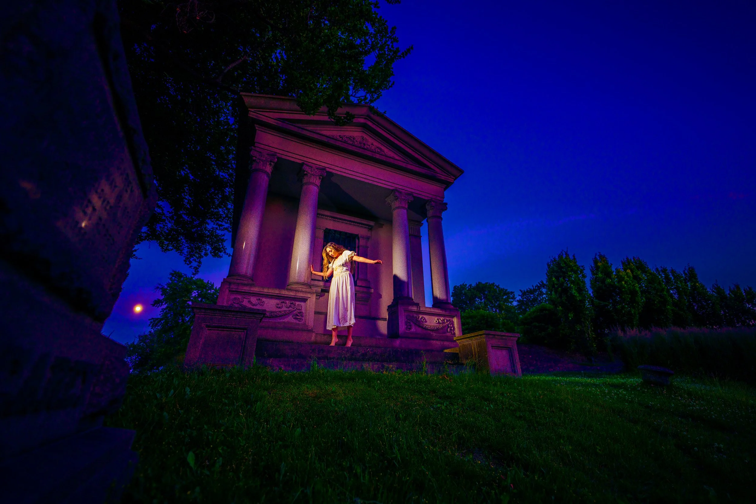 A woman in a white dress standing on steps in front of a classical building with columns, illuminated by purple and pink lighting, at dusk.